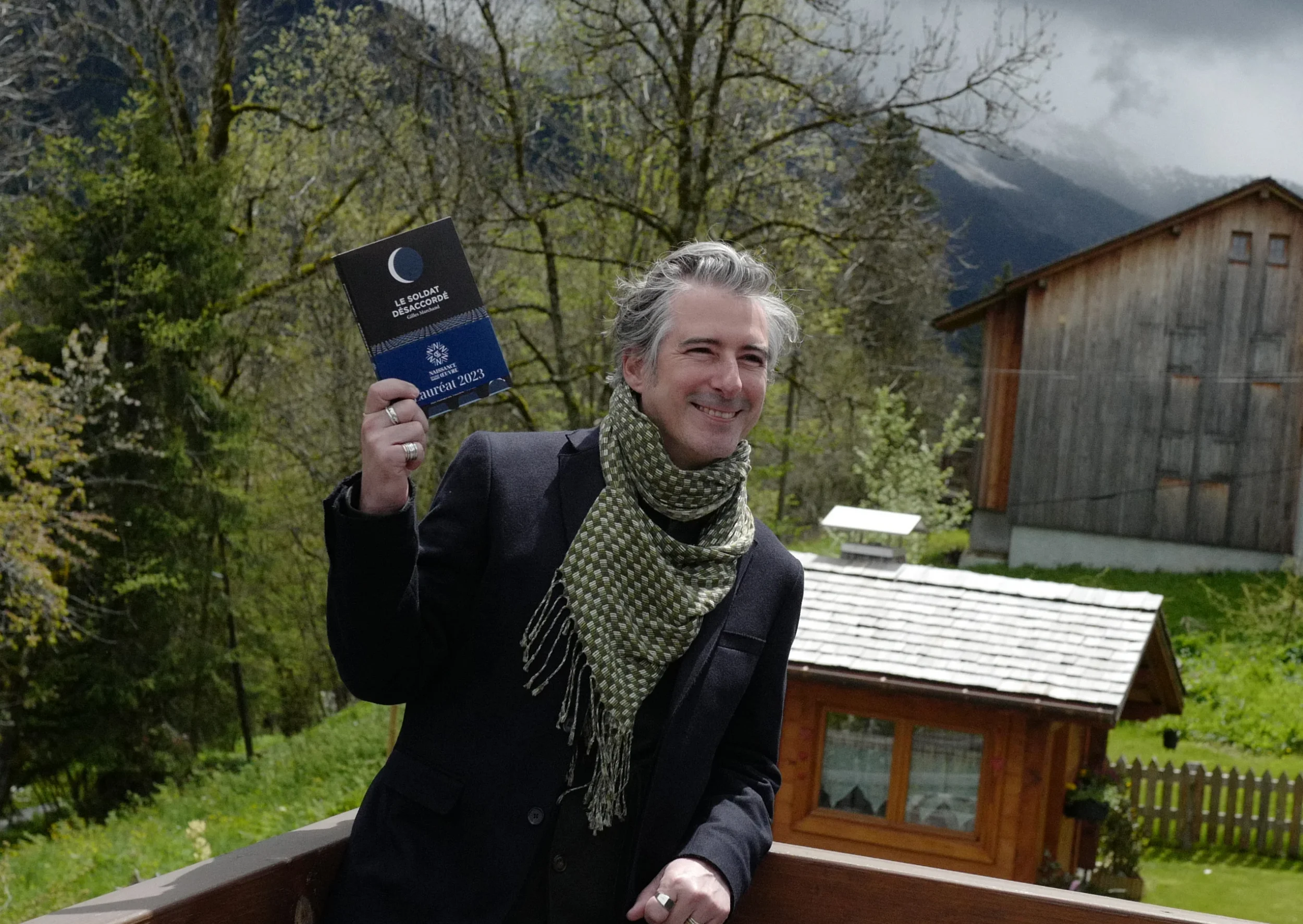 L'auteur souriant tenant un livre intitulé 'Le Soldat Désaccordé', avec un paysage montagneux en arrière-plan.