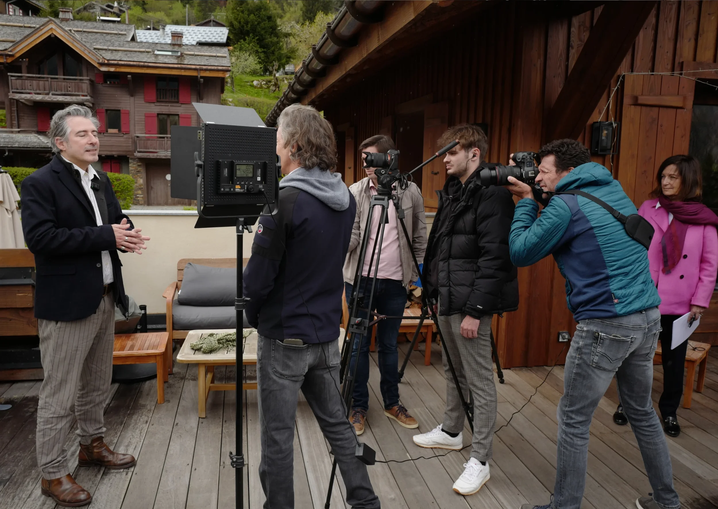 Une personne parle devant un groupe de journalistes avec des appareils photo et des caméras. Le groupe écoute attentivement à l'extérieur, avec des maisons en bois en arrière-plan.
