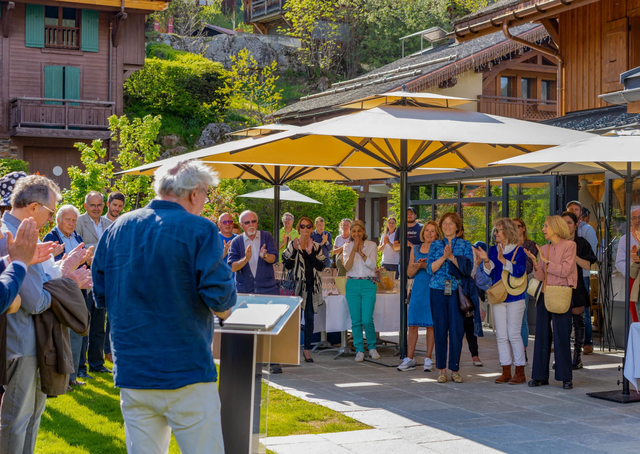Groupe de personnes assises et debout lors d'un rassemblement en plein air, sous des parasols, devant des maisons en bois dans un environnement verdoyant.