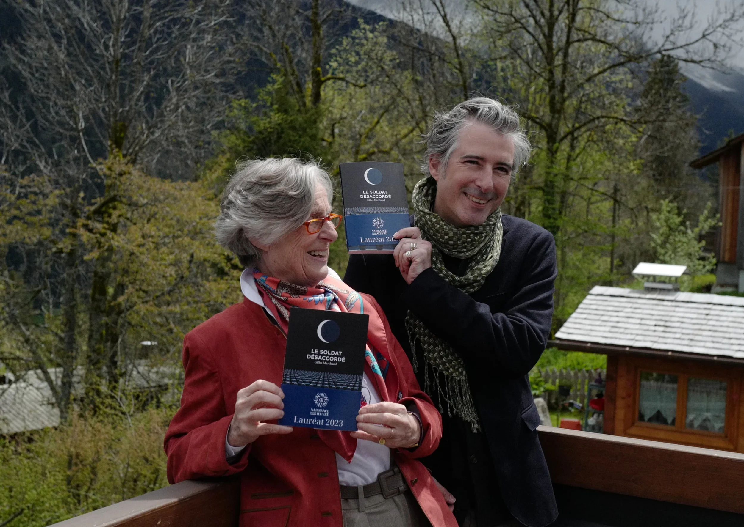 Deux personnes portent des prix de livre, une femme âgée avec des lunettes rouges et un homme avec une écharpe à carreaux, dans un environnement extérieur avec des arbres et des montagnes en arrière-plan.