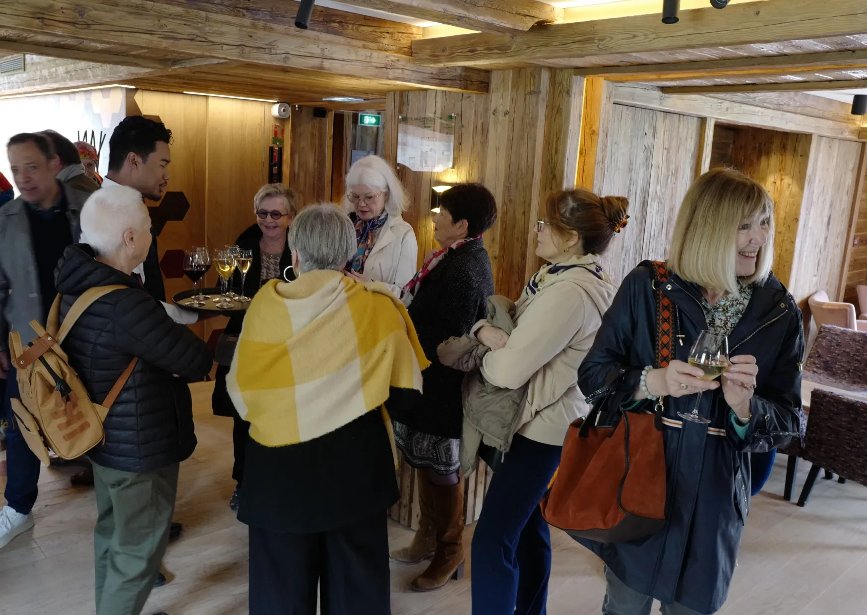 Groupe de personnes, principalement des femmes, socialisant dans un espace intérieur en bois, certaines avec des verres de vin ou de l'eau.