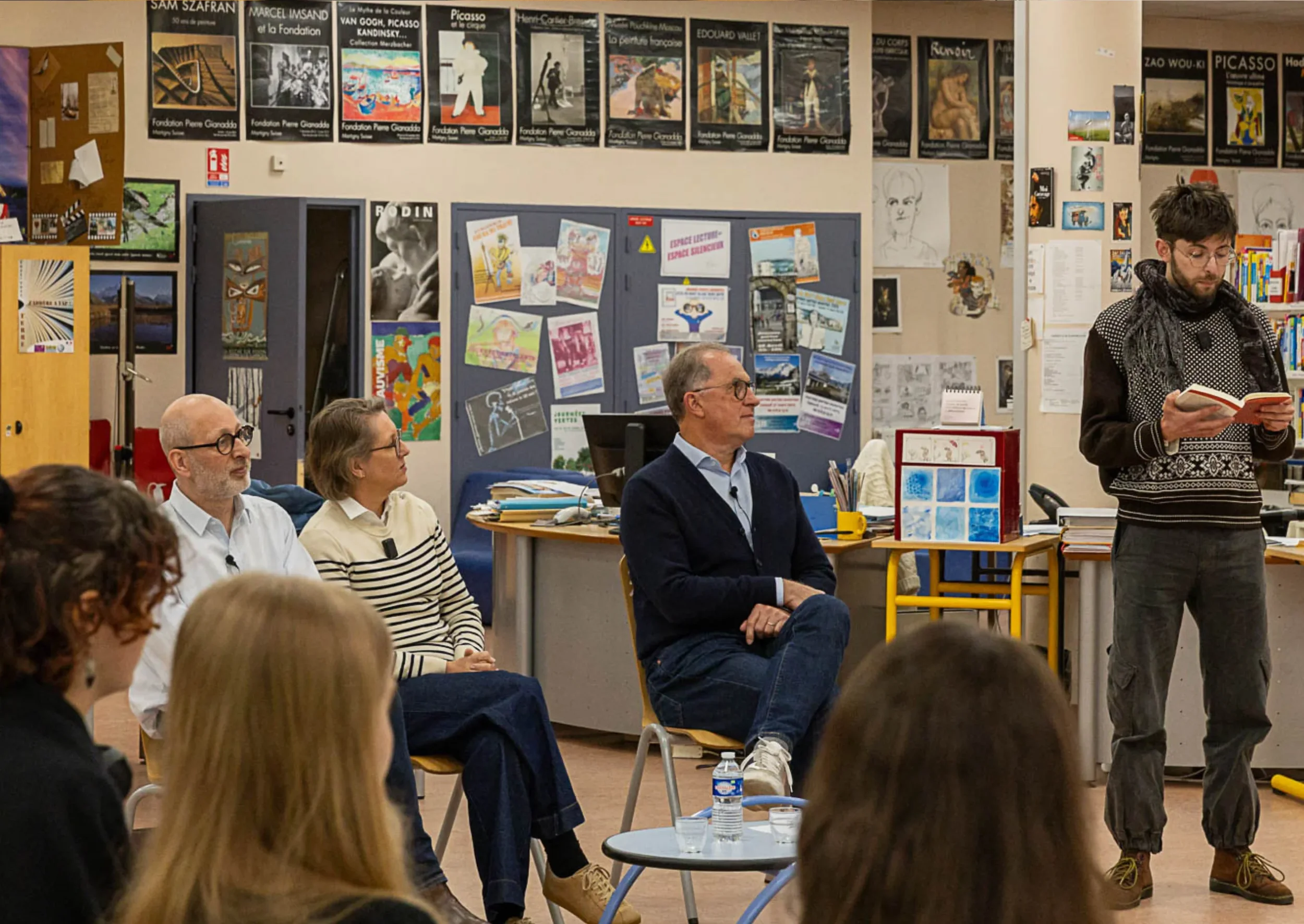 Une scène intérieure d'une salle de classe ou d'une réunion avec plusieurs personnes assises et une personne debout qui lit un livre. Le mur est décoré avec plusieurs affiches, posters d'art et dessins. Il y a des tables avec des documents, un ordina