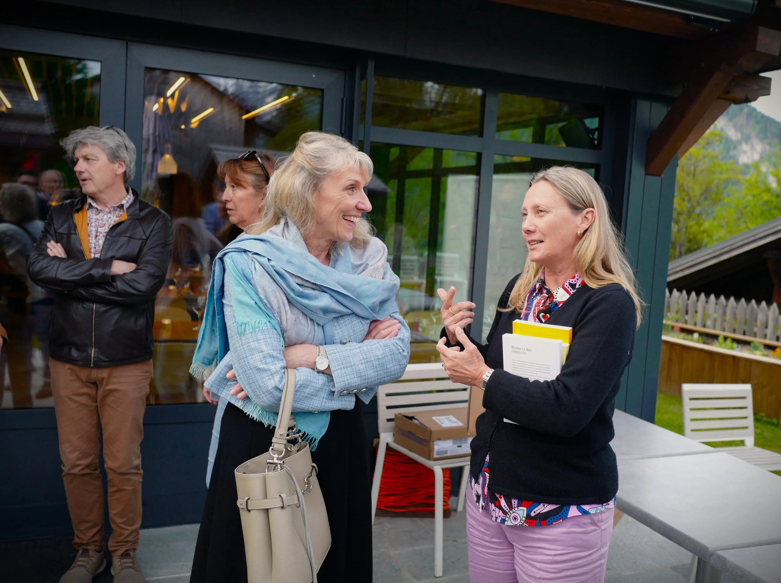 Trois femmes discutent devant une vitrine lors d'un événement en plein air. L'une d'elles, avec des cheveux gris, porte une écharpe bleue et un sac beige, et l'autre, avec des cheveux blondes, tient un livret ou un livre. Un homme avec des cheveux gr