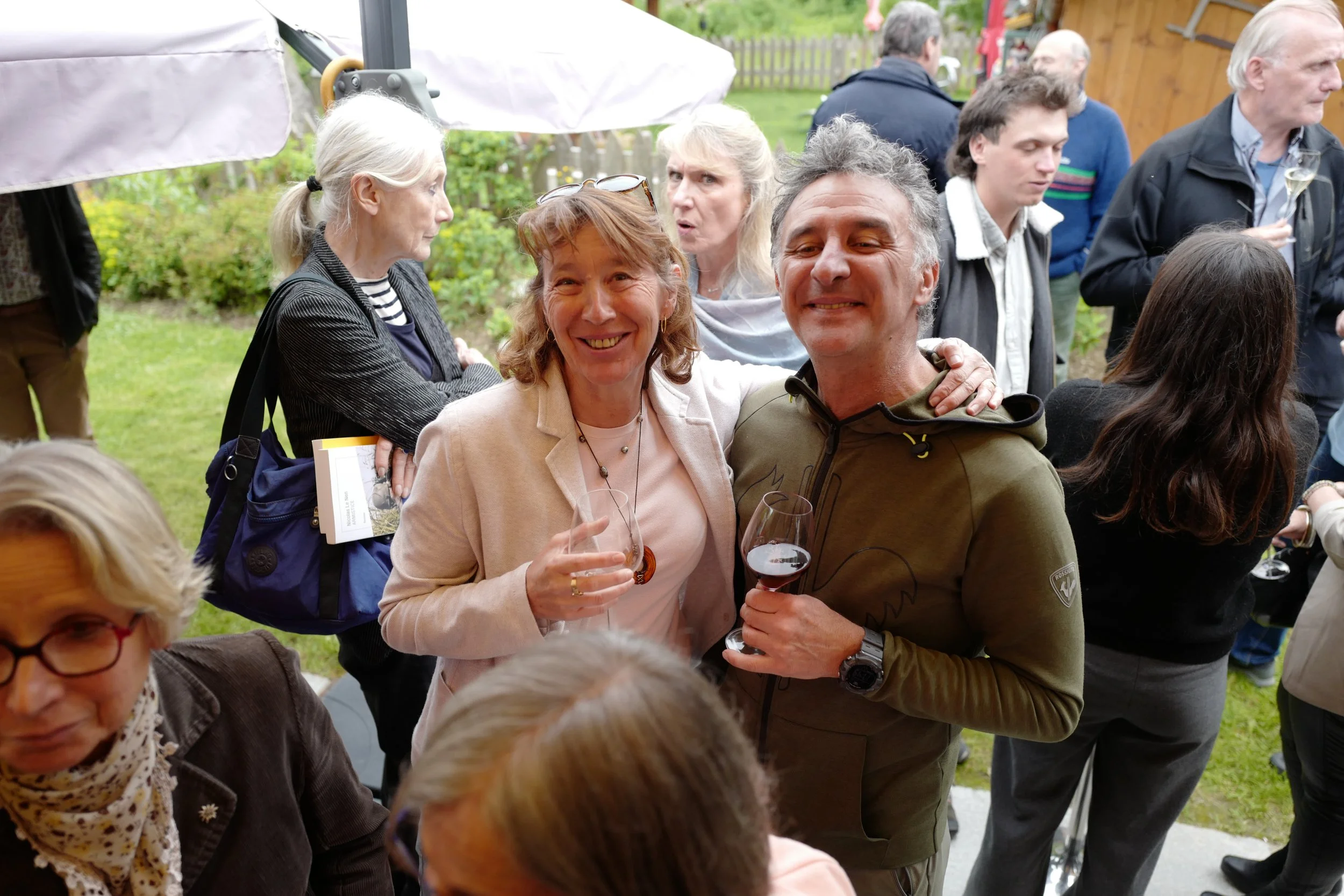 Groupe de personnes lors d'une réunion ou fête en extérieur, plusieurs tiennent un verre de vin et discutent.
