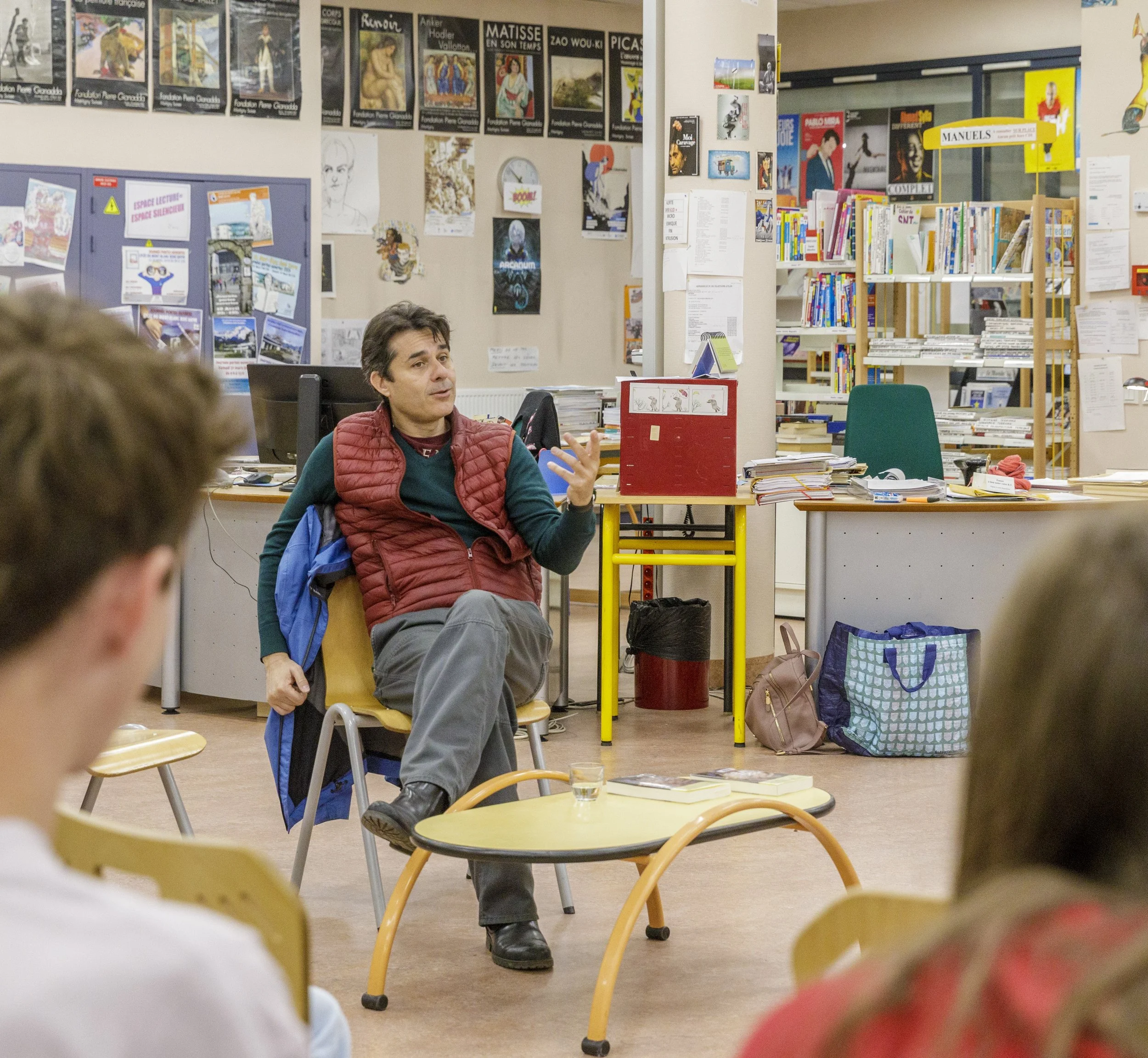Une salle de bibliothèque ou salle de classe avec des étagères remplies de livres, un tableau d'affichage, et un homme assis en discussion, avec plusieurs personnes en présence dont deux enfants en premier plan.