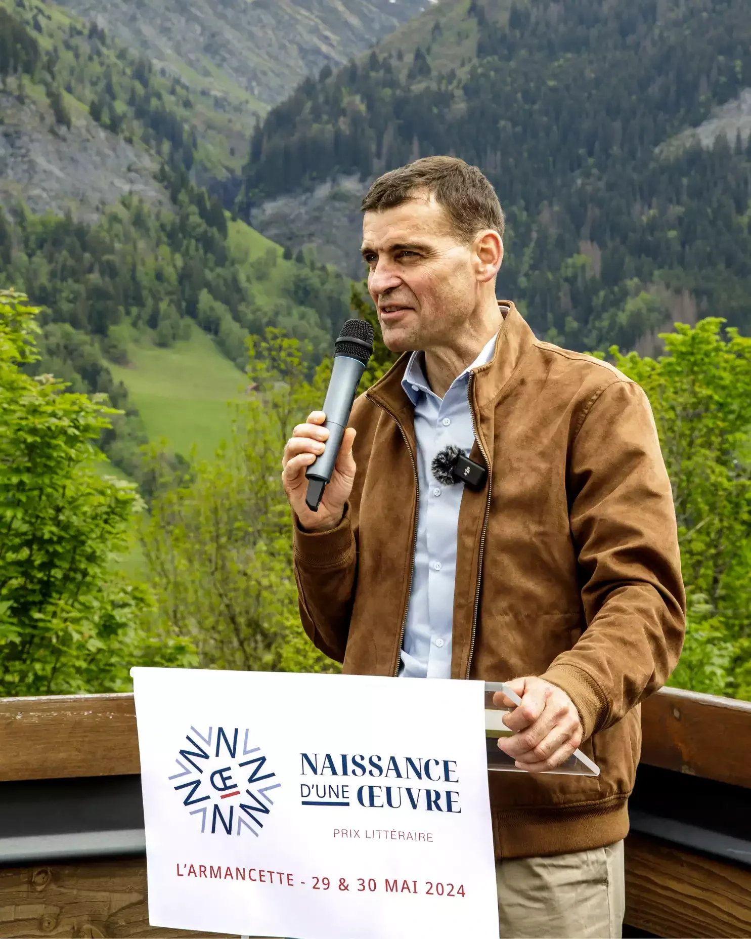 Un homme tenant un micro lors d'une présentation en plein air dans un environnement montagneux, avec un panneau indiquant un prix littéraire aura lieu les 29 et 30 mai 2024 à L'Armacette.