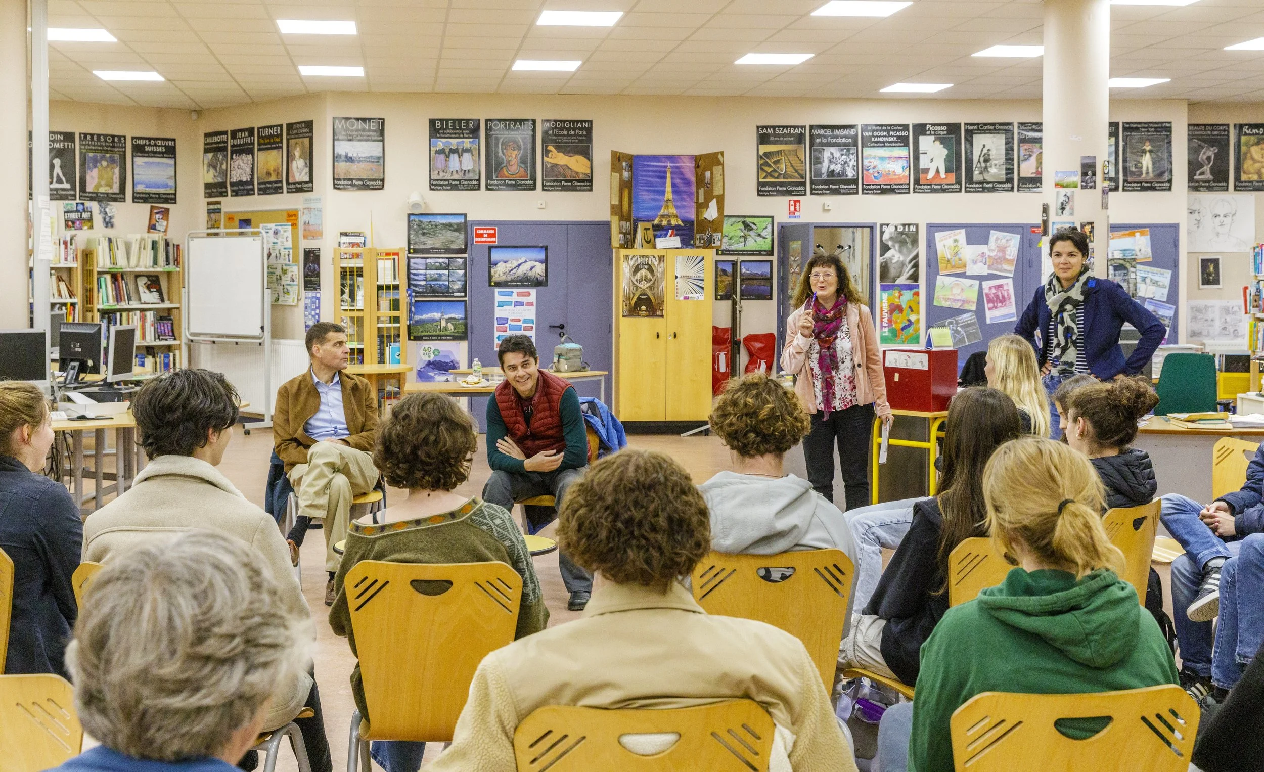 Une scène dans une bibliothèque ou un centre communautaire où plusieurs personnes participent à une discussion ou un atelier. Deux femmes se tiennent devant le groupe, une parlant et l'autre écoutant. Des affiches et des œuvres d'art sont visibles au