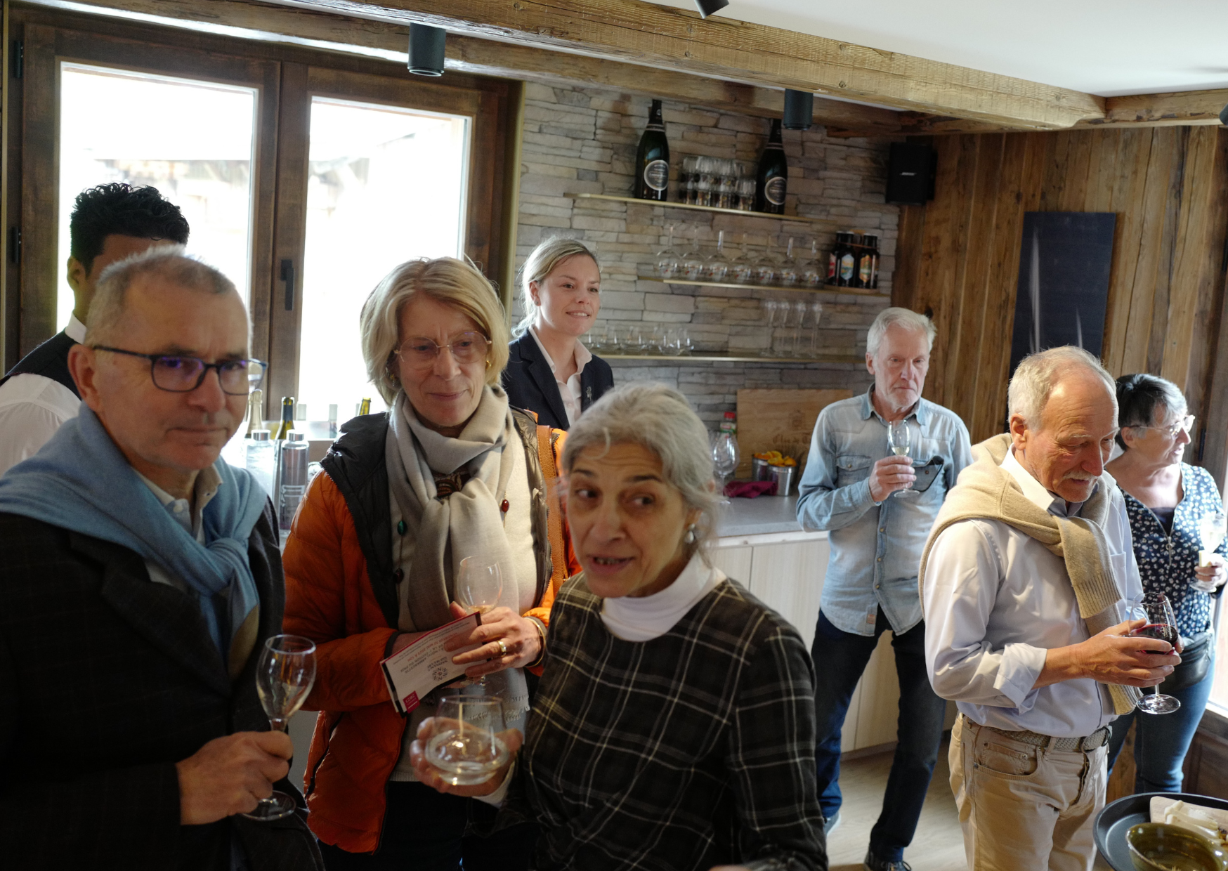 Groupe de personnes à une réception ou un événement, tenant des verres de vin, dans une pièce au décor en bois avec fenêtres naturelles en arrière-plan.