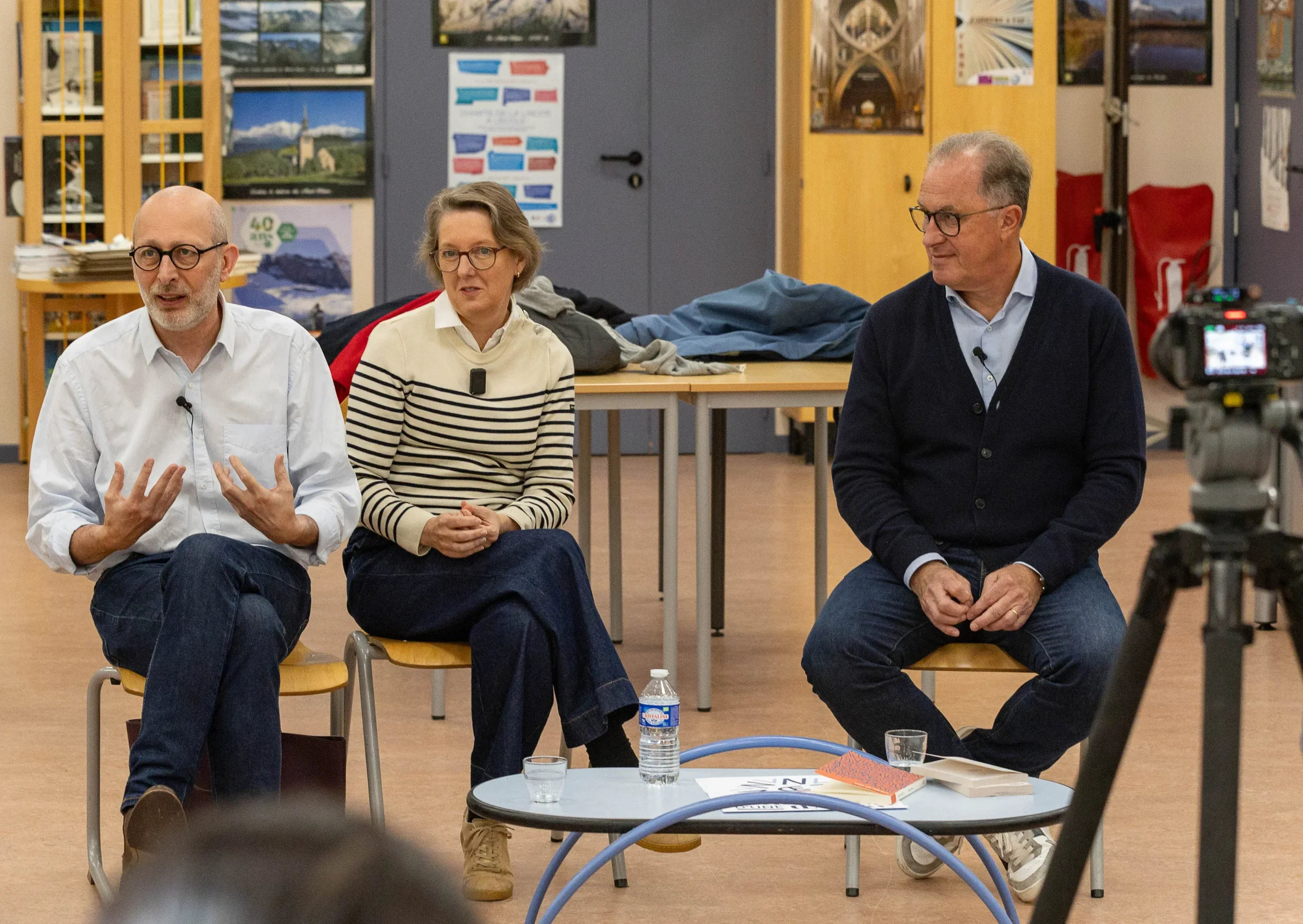 Trois personnes assises lors d'une discussion ou d'une interview, deux hommes et une femme, dans une salle avec des affiches et des livres en arrière-plan, une table avec une bouteille d'eau, des verres et des documents devant eux.