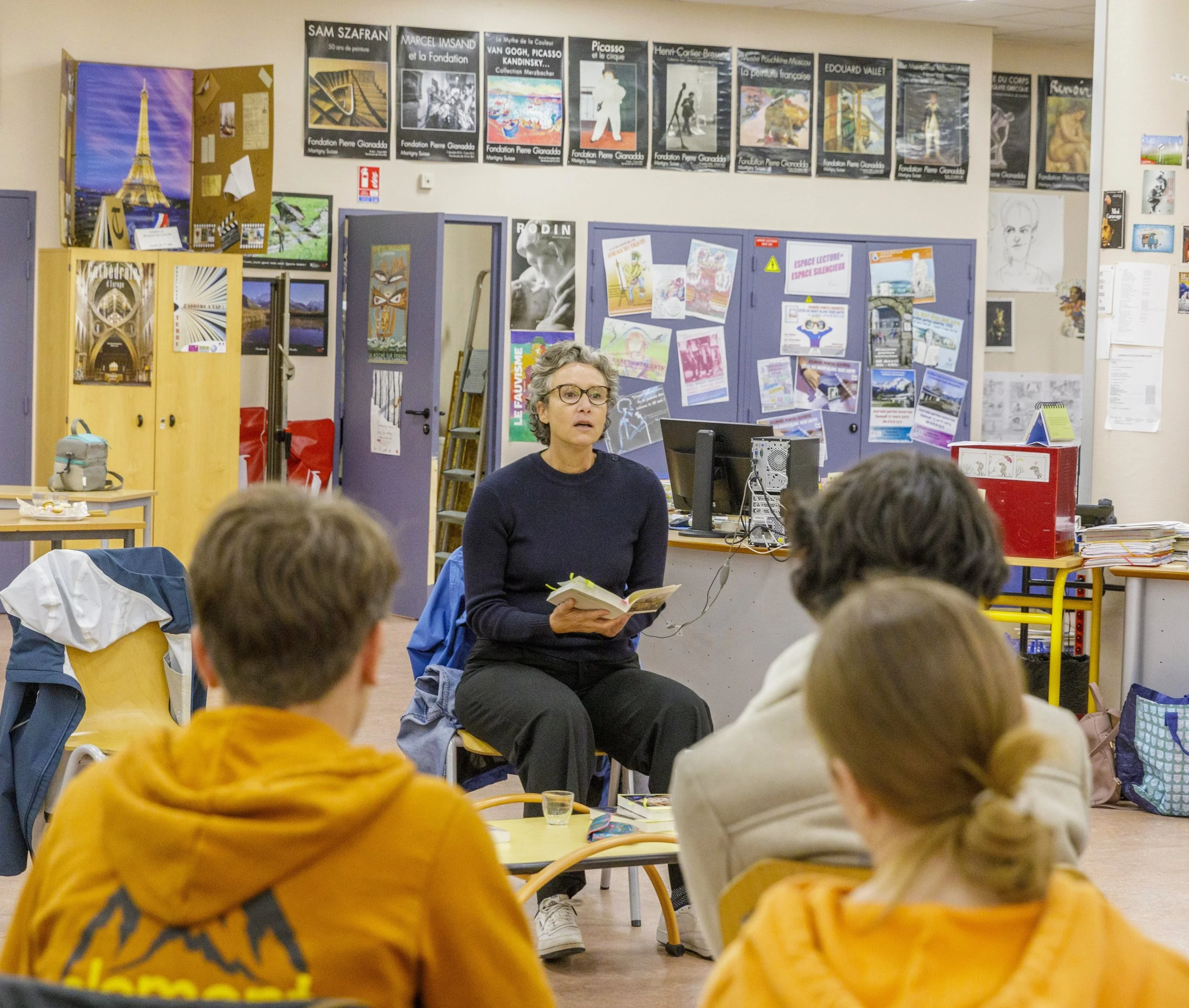 Une femme avec des lunettes parle devant un groupe d'étudiants dans une salle de classe, avec de nombreux posters et affiches sur les murs et des étagères contenant des livres et des objets.