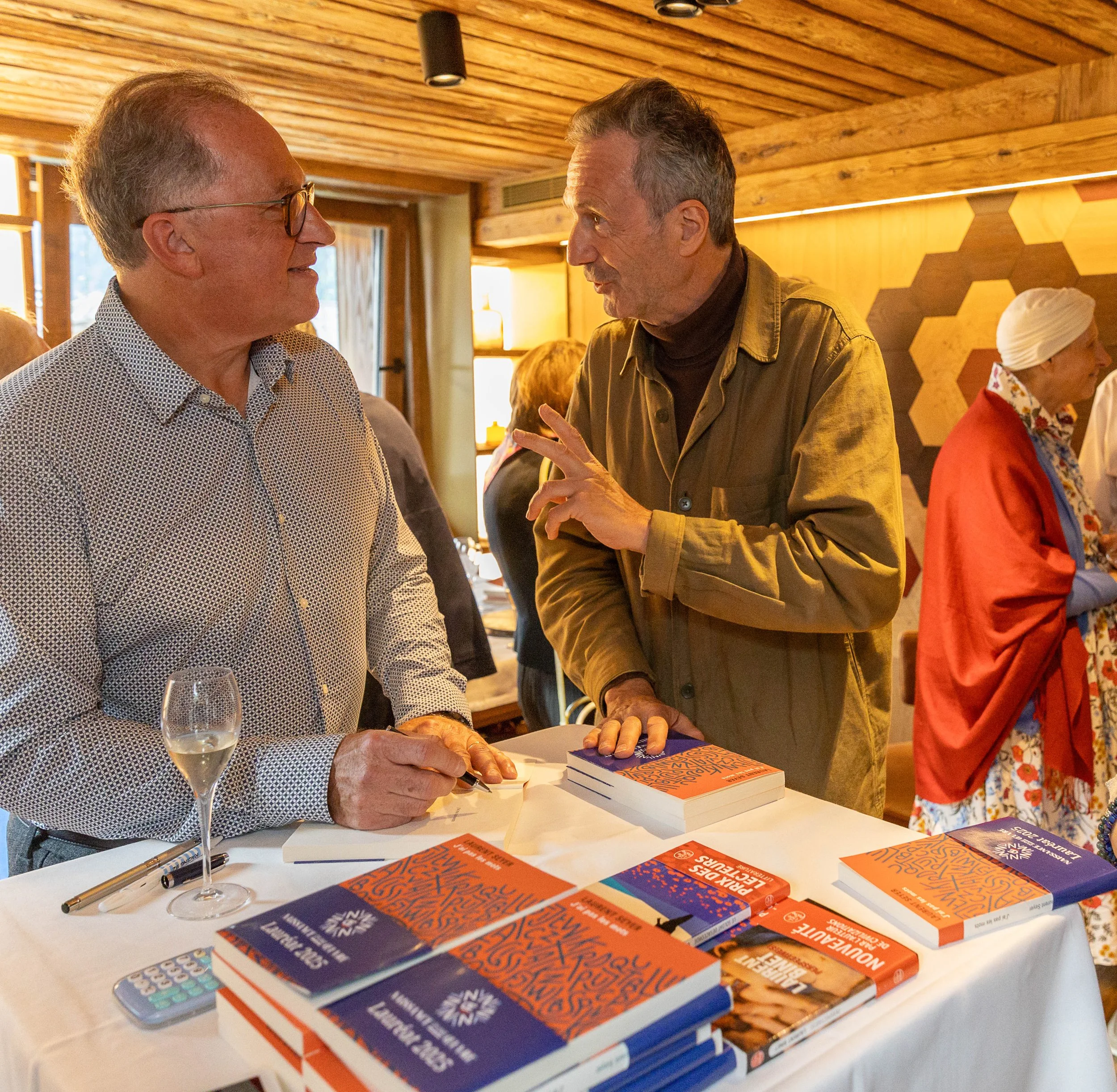 Deux hommes discutent dans une pièce avec un mur en bois, devant une table avec plusieurs livres ou brochures orange et bleus, un verre de vin blanc, un stylo et un téléphone portable.