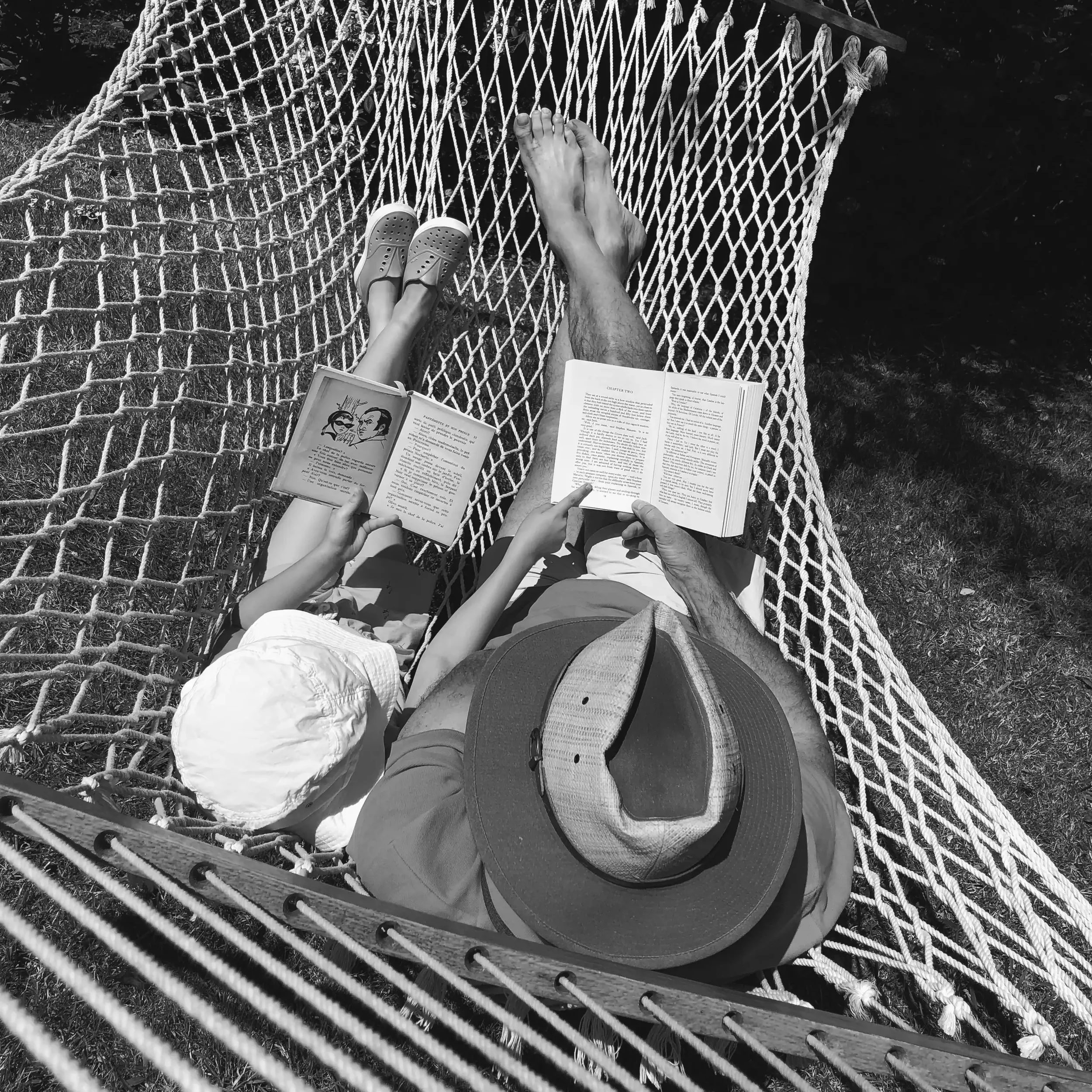 Personne allongée sur un hamac en train de lire un livre, vue de haut, en noir et blanc, avec des chaussures de sport visibles et un livre ouvert.