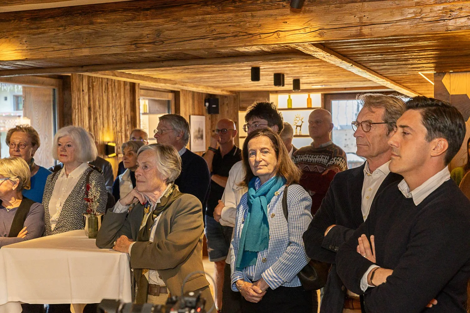 Un groupe de personnes assises et debout lors d'une réunion ou d'un événement dans une pièce en bois, écoutant attentivement.