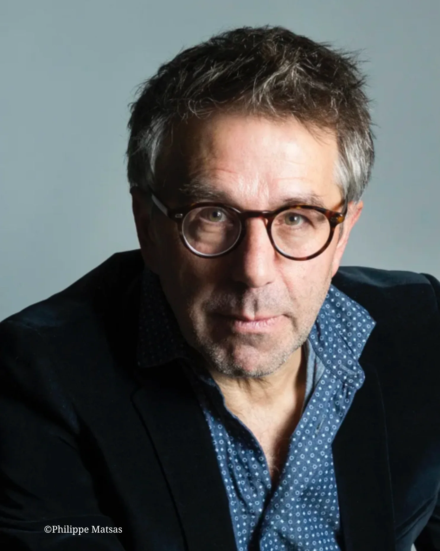 Portrait d'un homme portant des lunettes, habillé d'une chemise à pois et d'une veste, avec des cheveux bruns en désordre et un léger sourire.