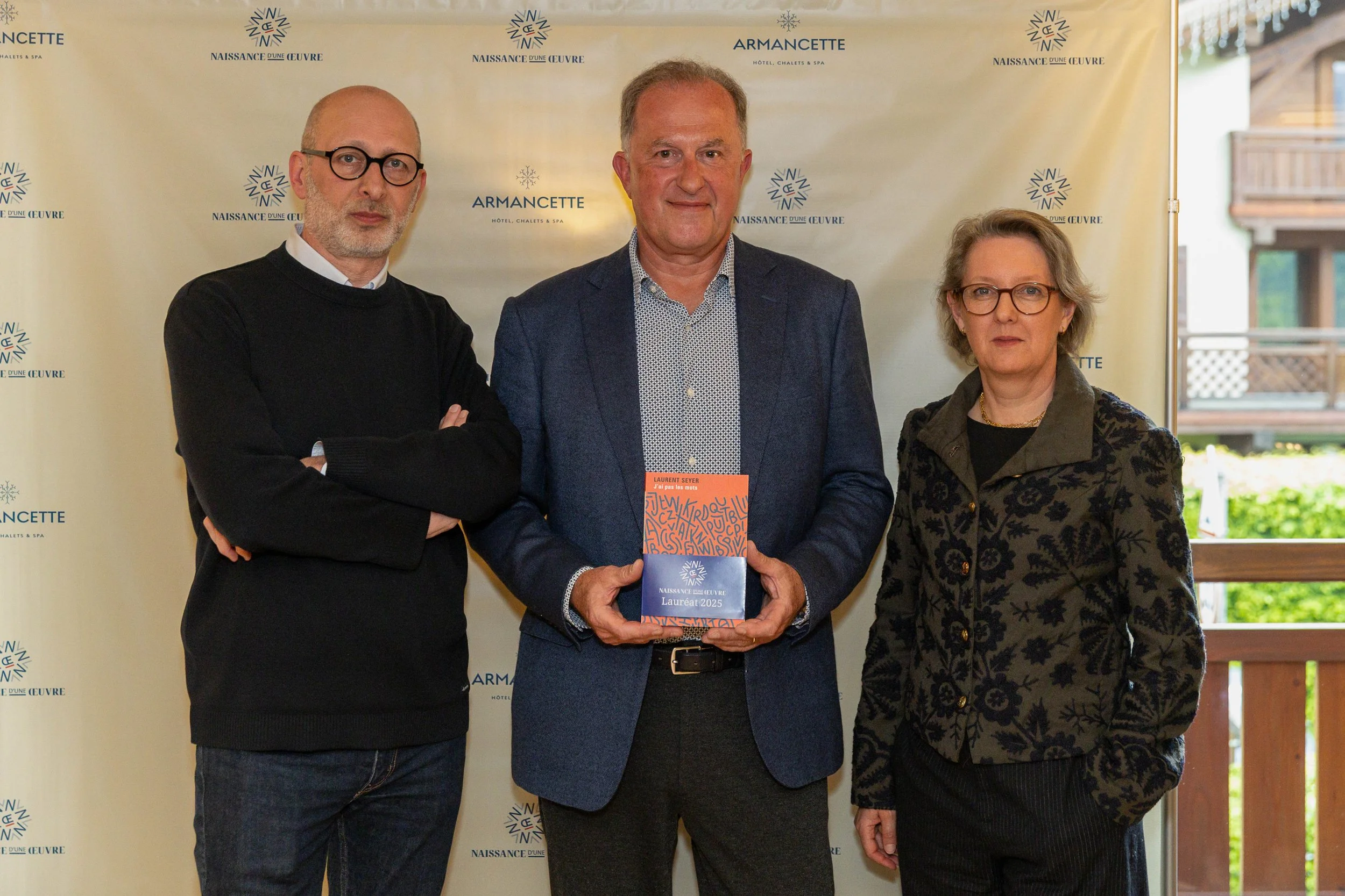 Trois personnes posent pour une photo lors d'un événement, avec un arrière-plan contenant des logos et noms liés à un hôtel ou spa nommé Armancette, avec un homme au centre tenant un livre ou un prix, et deux autres personnes à ses côtés.