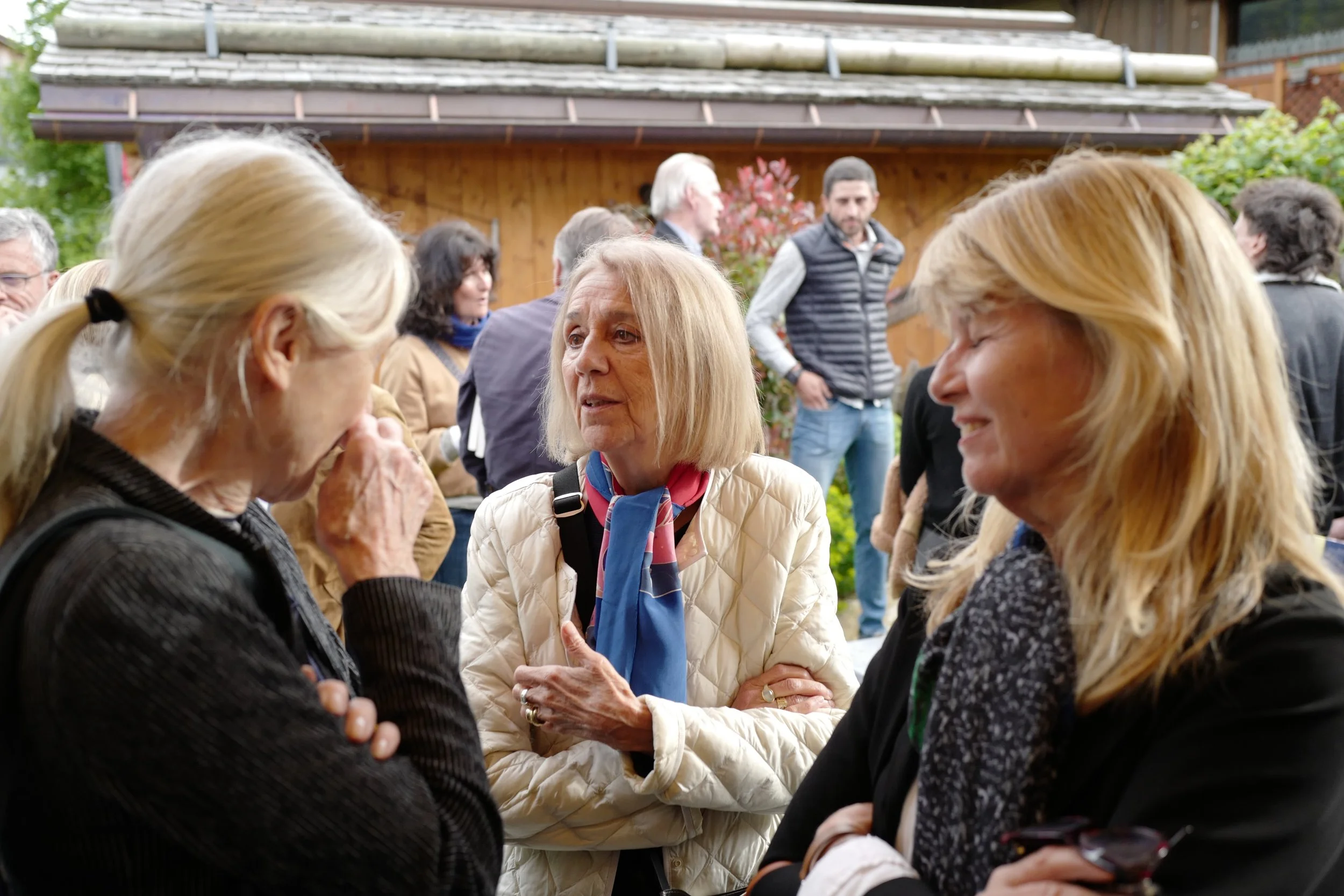 Groupe de personnes âgées parlant et se tenant par les bras lors d'une rencontre dans un jardin, avec des autres personnes en arrière-plan.