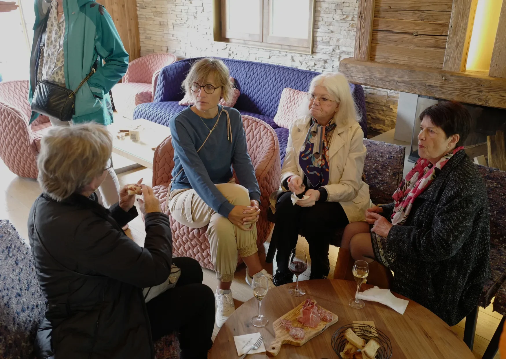 Groupe de cinq femmes âgées assises et parlant dans un salon, avec une table basse contenant du vin, du pain et des charcuteries.