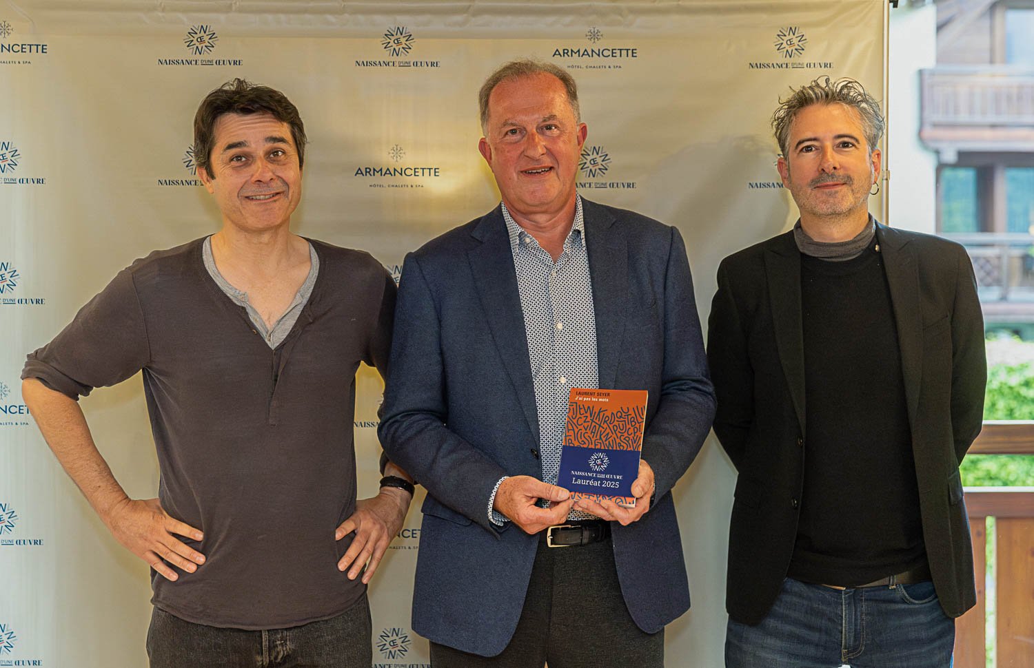 Trois hommes posant pour une photo lors d'une cérémonie de remise de prix, avec un fond à motifs et le logo "Naissance d'une Œuvre".