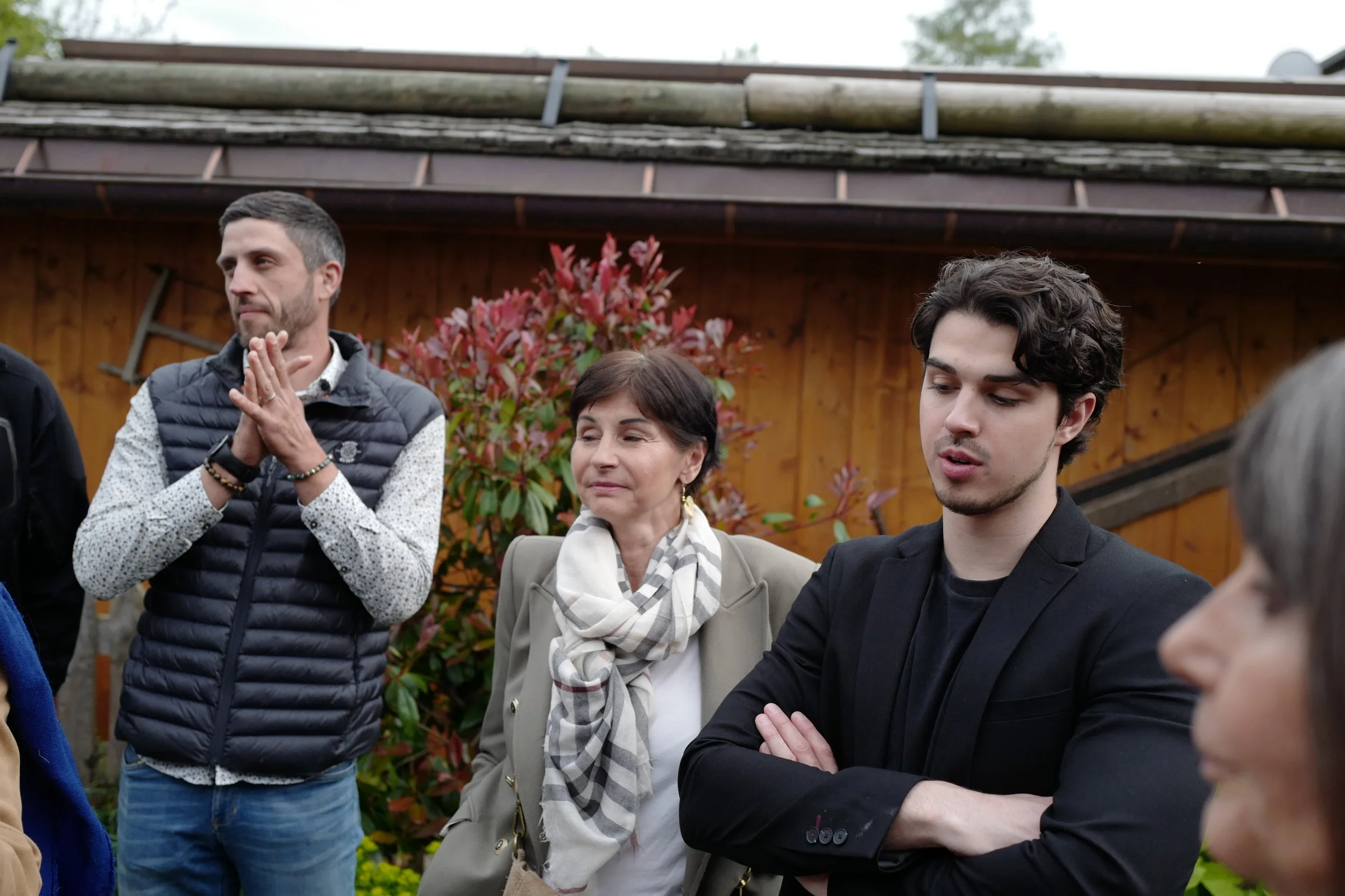 Groupe de personnes en discussion à l'extérieur, avec un bâtiment en bois en arrière-plan et un arbre avec des feuilles rouges.
