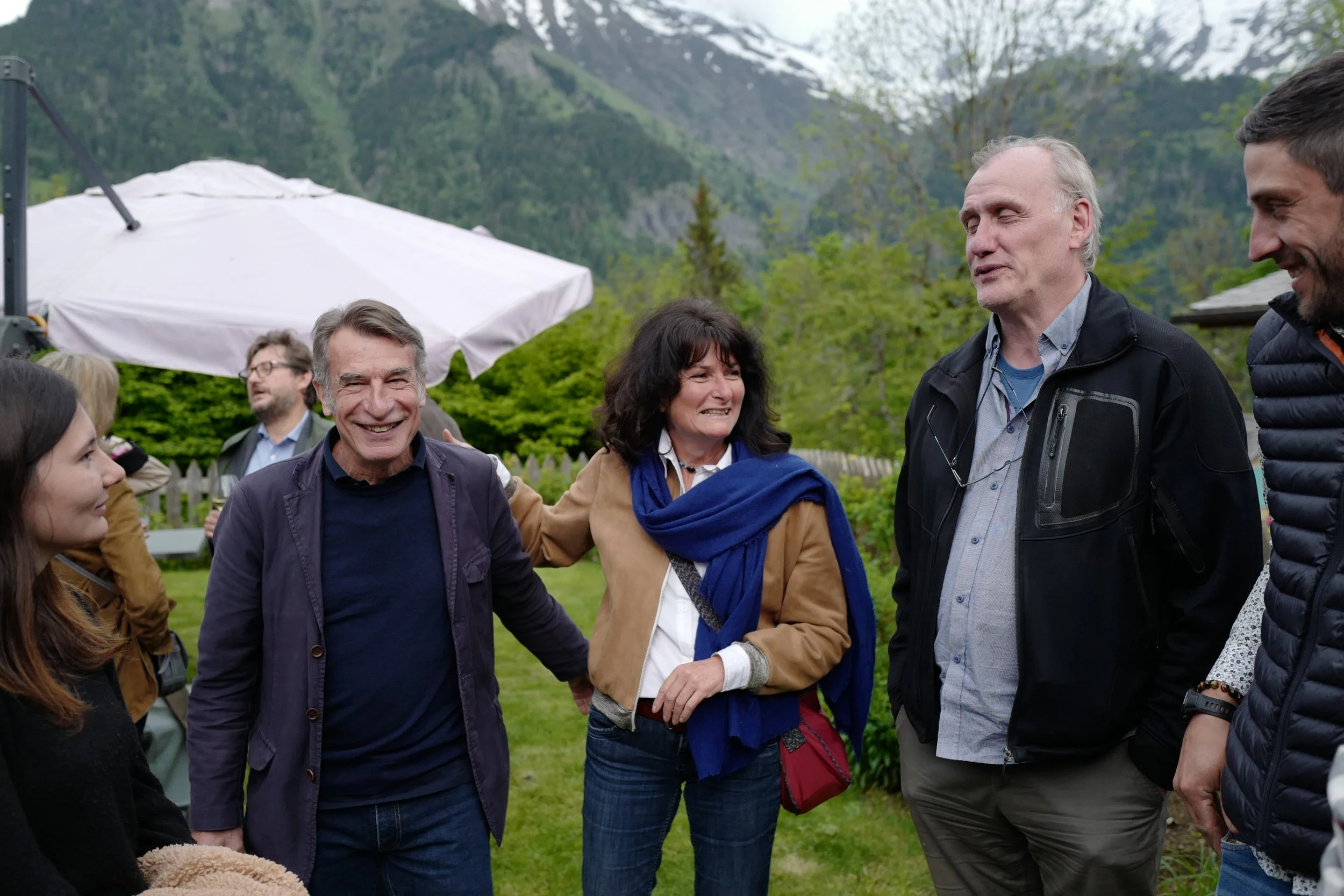 Groupe de personnes discutant et souriant à l'extérieur, avec des montagnes et un ciel nuageux en arrière-plan.