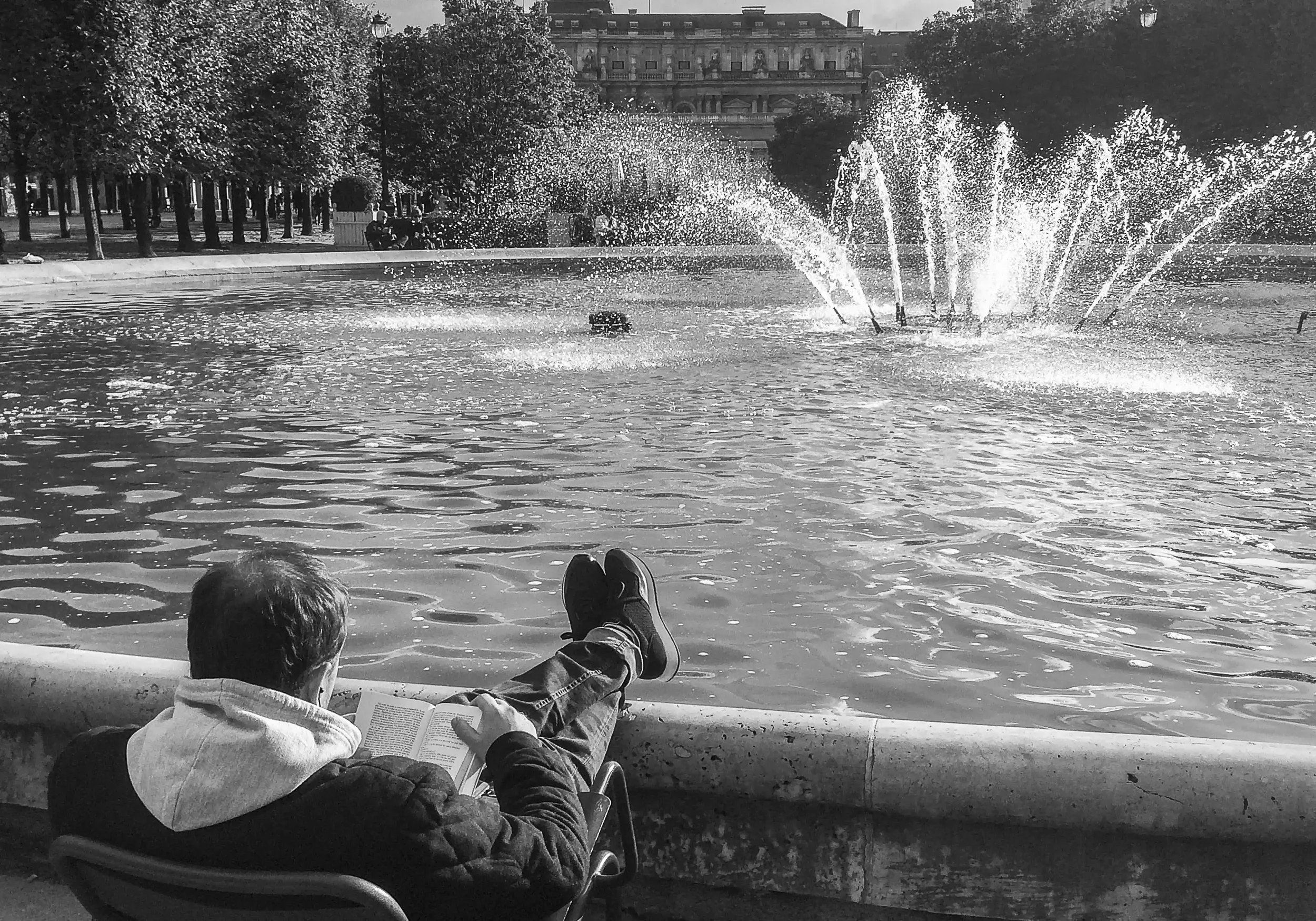 Deux enfants assis sur un bord de fontaine dans un parc, en train de lire un livre, avec un bâtiment historique en arrière-plan.