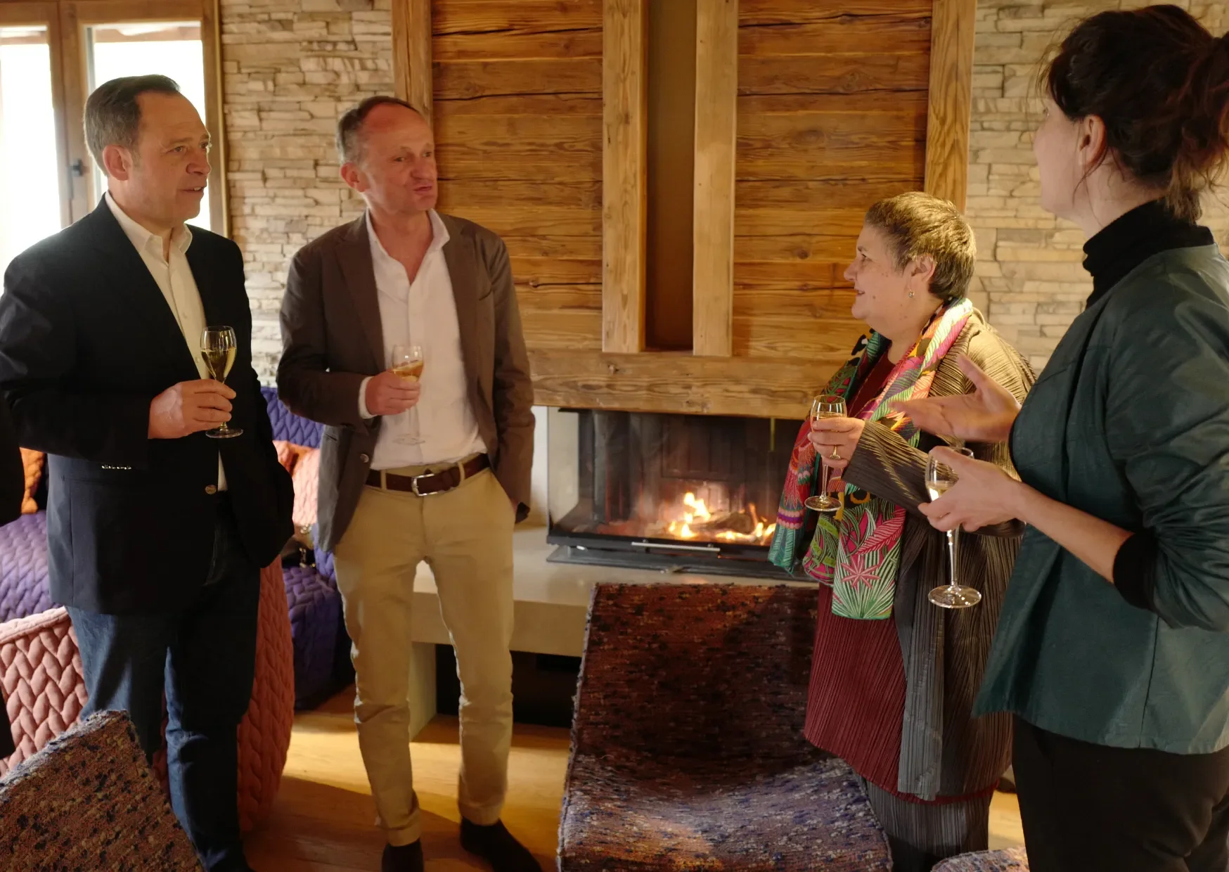 Groupe de cinq personnes discutant avec des verres de champagne, dans une pièce chaleureuse avec cheminée en pierre et mur en bois.
