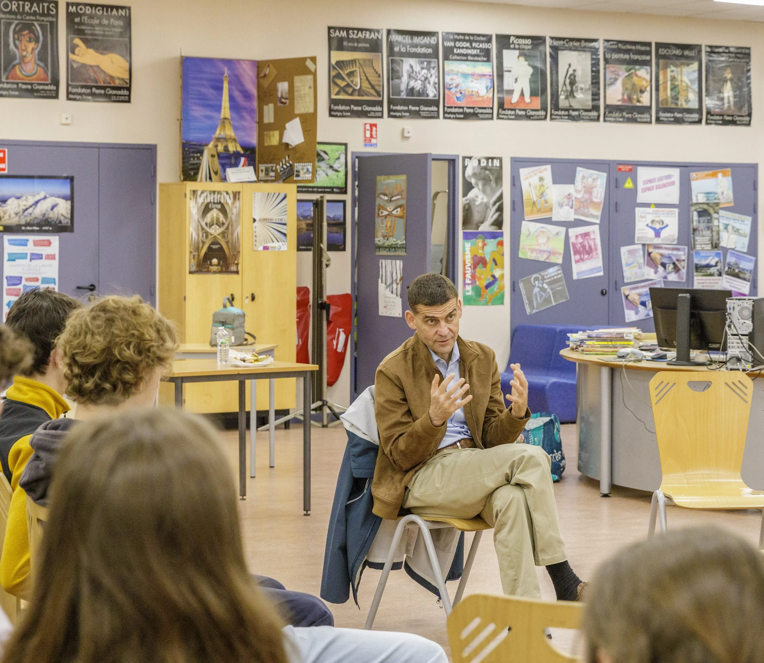 Un homme parle à un groupe d'élèves dans une salle de classe, avec des posters d'art et des images de la Tour Eiffel sur les murs.