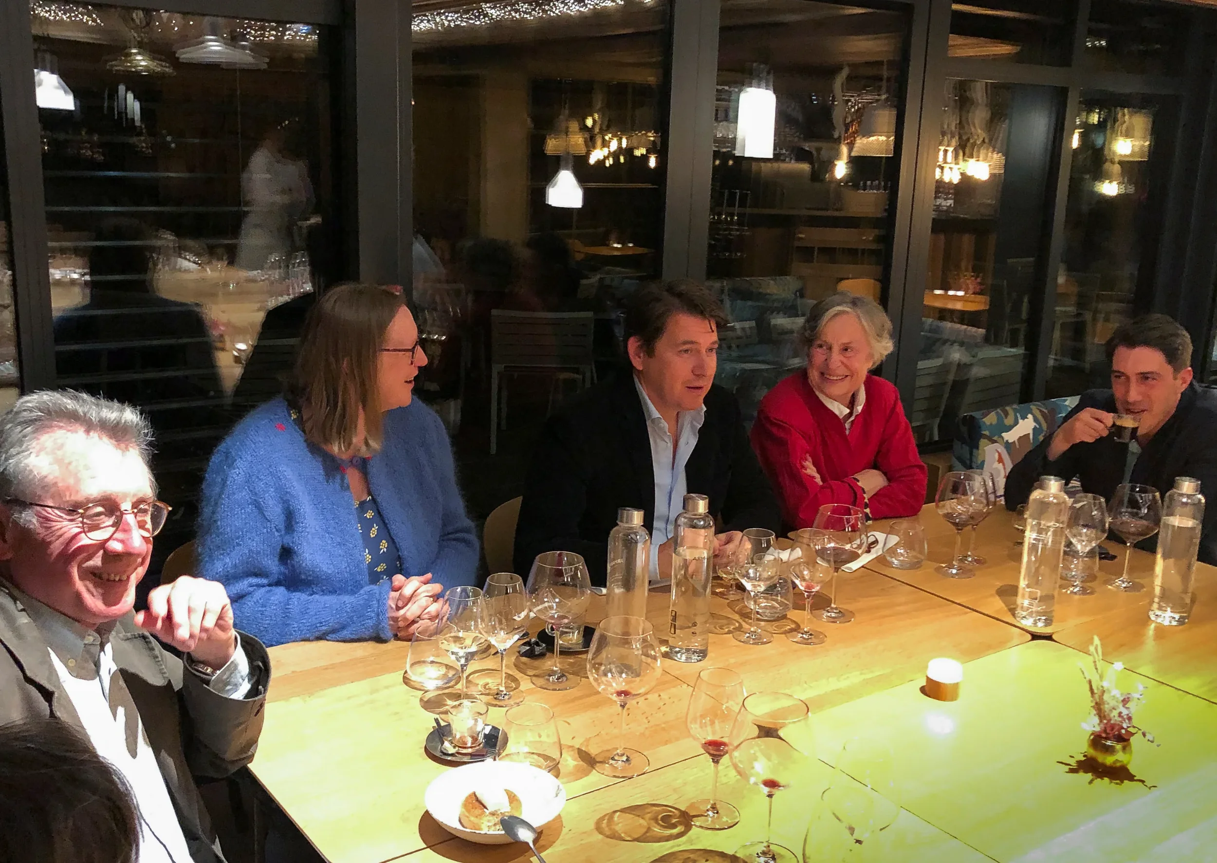 Groupe de personnes assises autour d'une table à dîner dans un restaurant, en train de parler et de sourire, ambiance chaleureuse avec éclairage tamisé.
