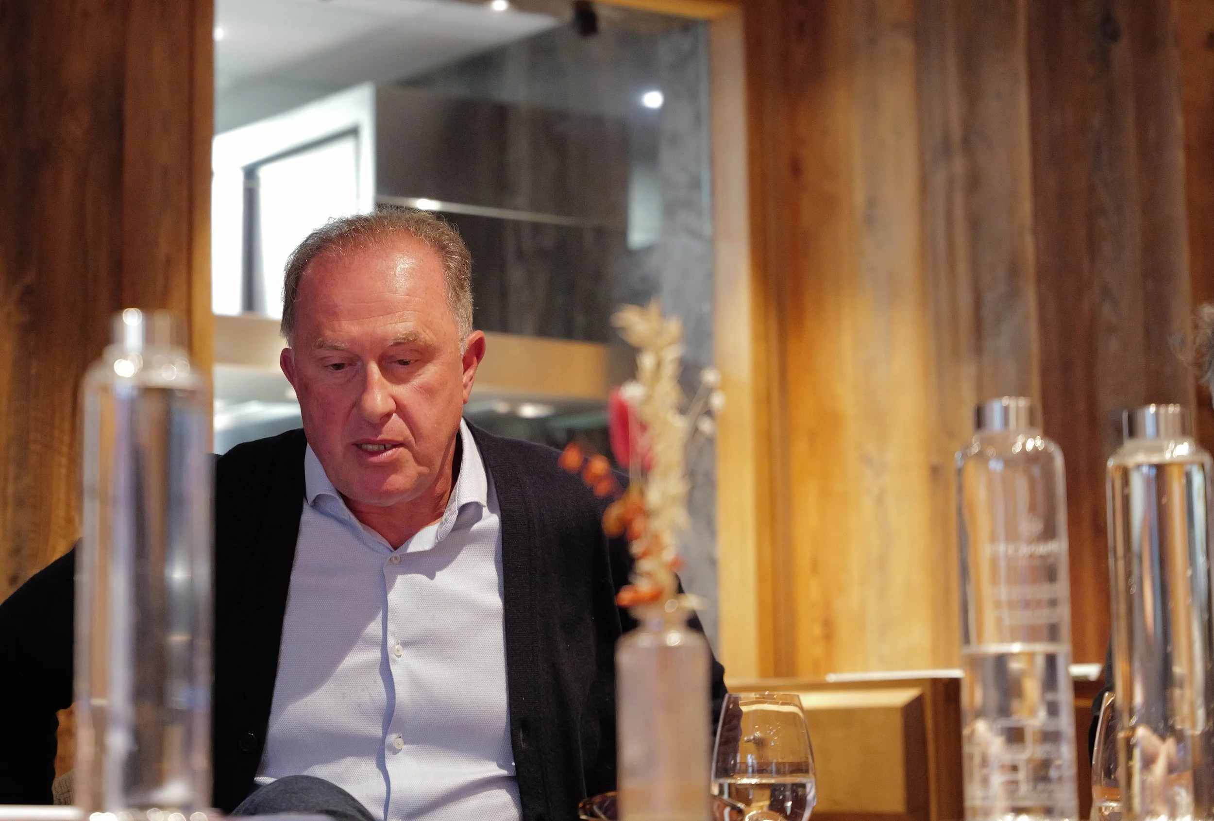 Un homme d'âge moyen avec une peau clair, des cheveux gris, portant une chemise blanche et un pull noir, assis dans un restaurant avec un décor en bois. Il semble être en train de parler ou de réfléchir.
