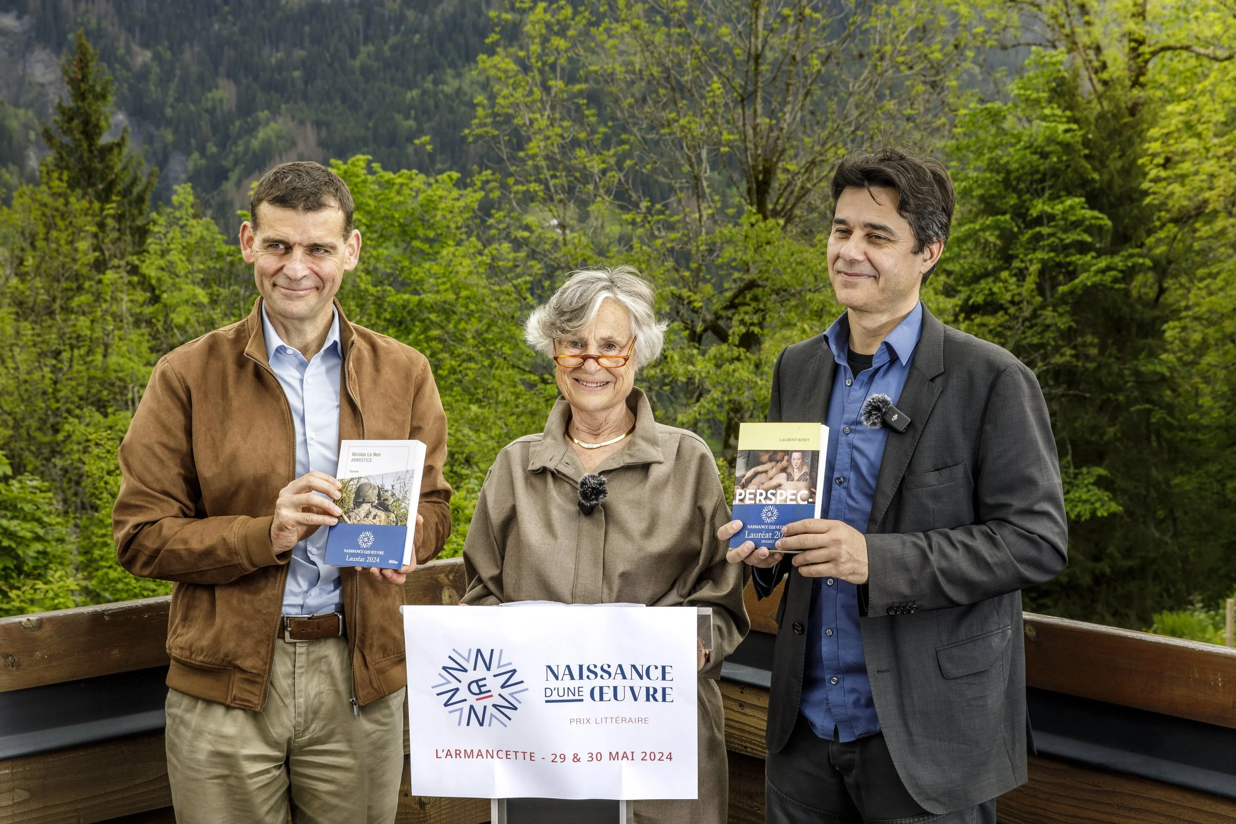 Trois personnes debout près d'une affiche promotionnelle, avec un paysage de forêt en arrière-plan. Ils tiennent des livres ou des brochures, suggérant un événement littéraire ou culturel.