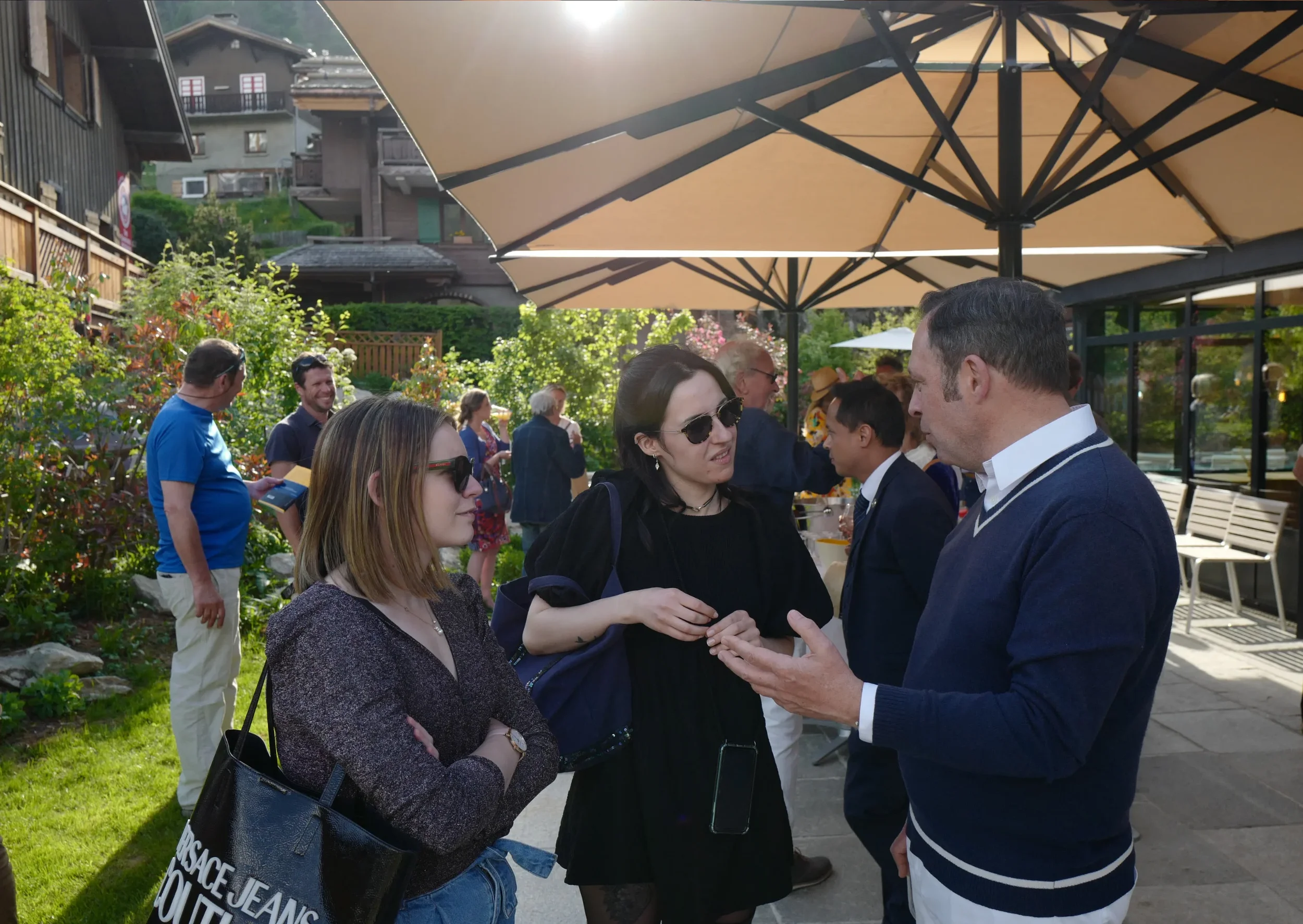 Groupe de personnes discutant et échangeant des salutations dans un jardin extérieur, sous de grands parasols beige, avec des maisons en bois en arrière-plan et une végétation luxuriante.