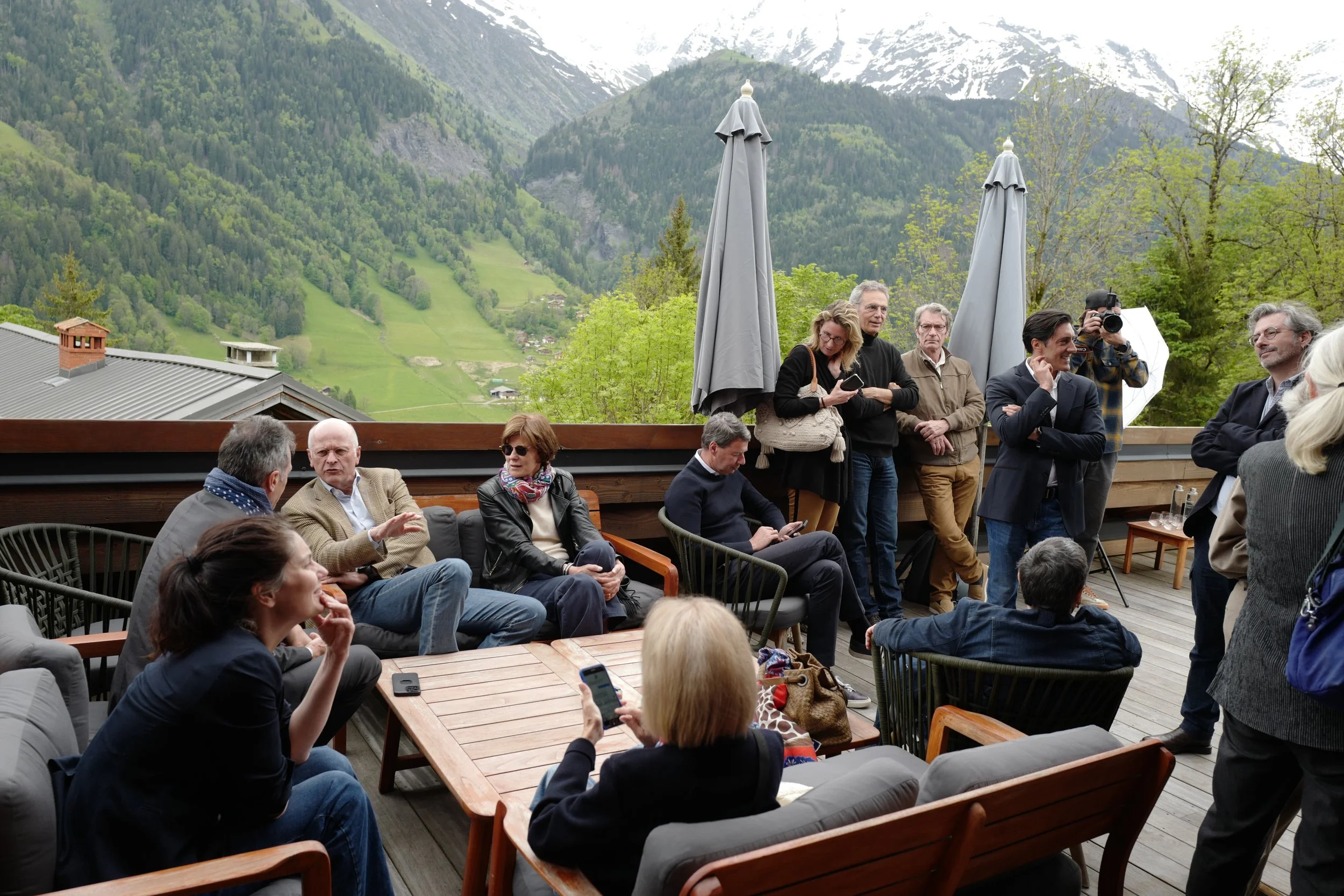 Groupe de personnes lors d'une réunion en plein air avec montagnes en toile de fond. Certaines personnes sont assises à une table, d'autres discutent debout, un homme photographie la scène, ambiance décontractée.