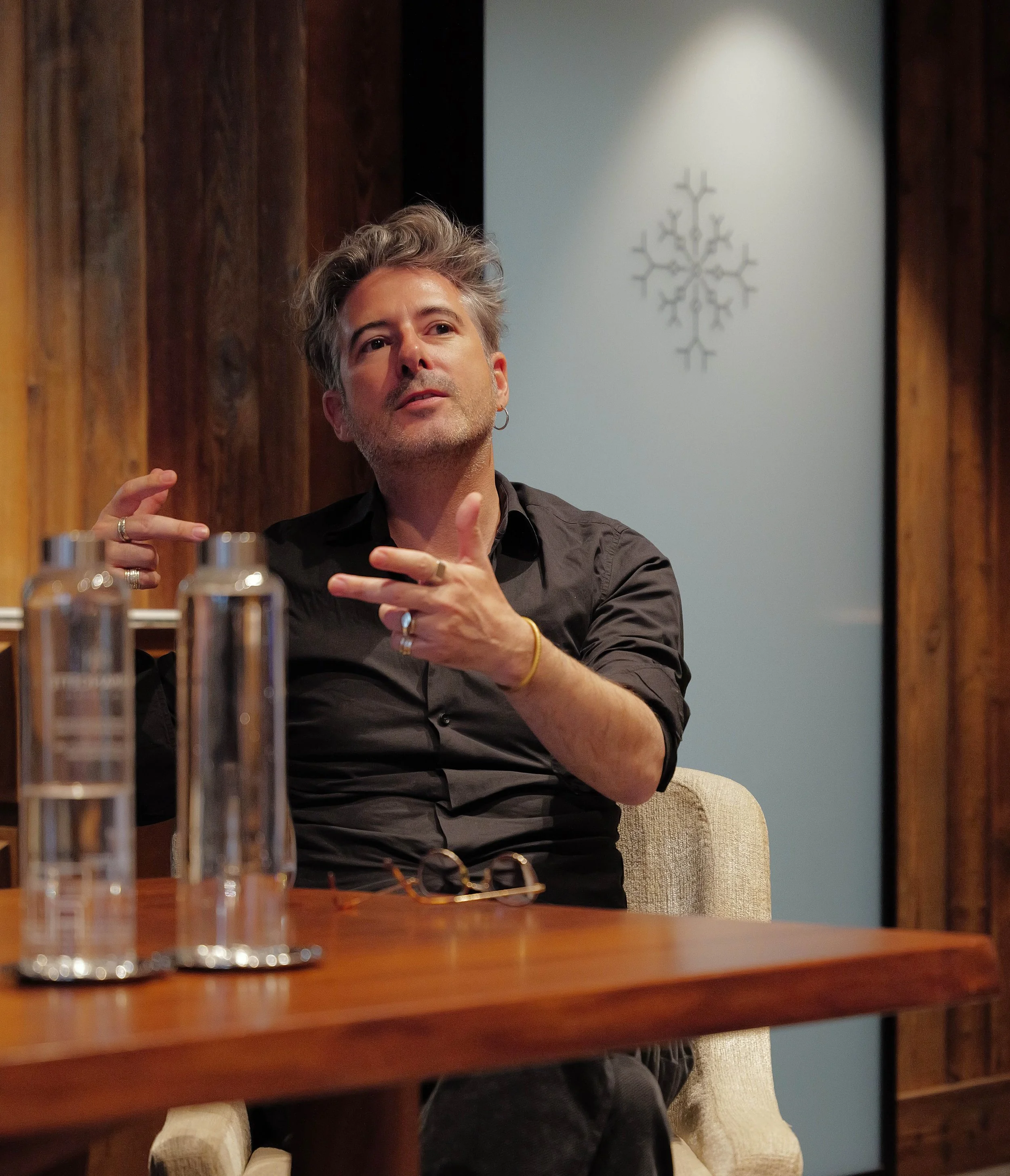 Un homme à cheveux gris en train de parler, assis à une table en bois, avec des verres en verre devant lui, dans une salle avec des murs en bois et un symbole en forme de flocon de neige sur le mur en arrière-plan.
