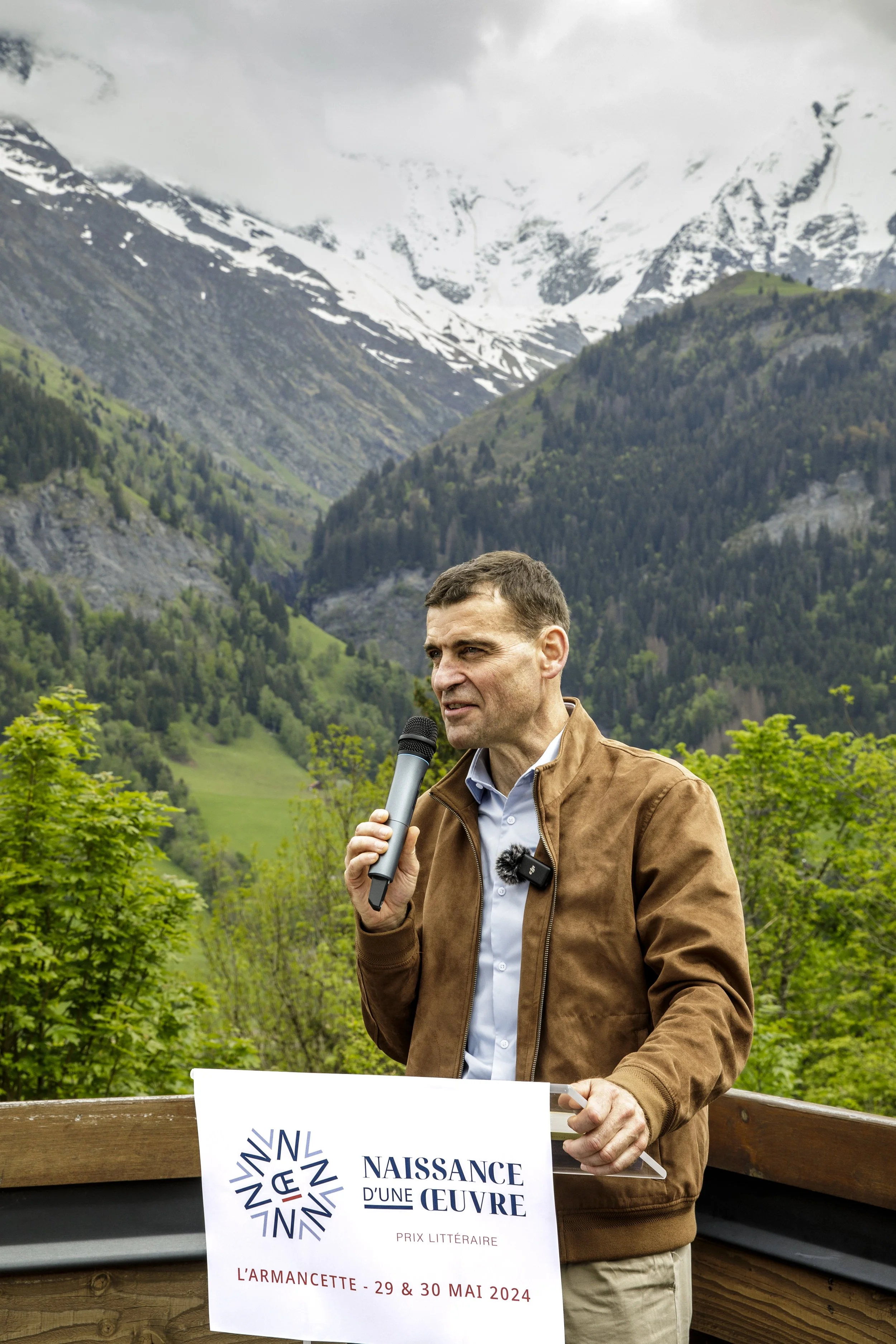 Homme parlant dans un micro lors d'un événement en plein air avec montagnes et forêt en arrière-plan, tenant une affiche pour un prix littéraire nommé 'Naissance d'une œuvre'.