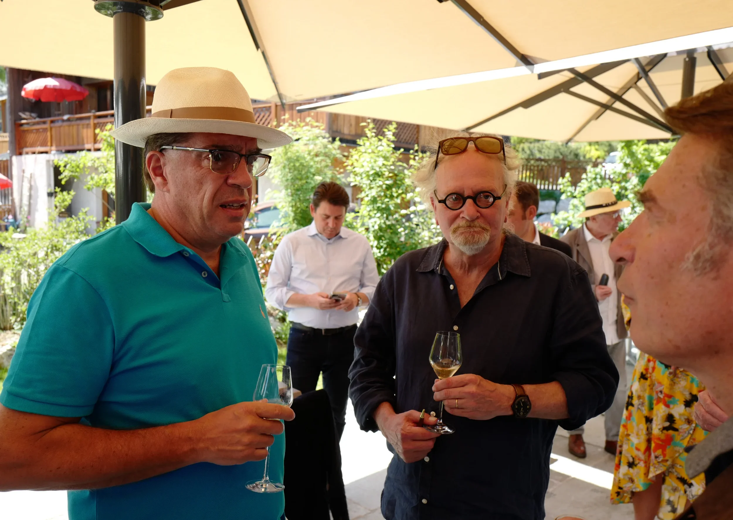 Groupe de personnes discutant lors d'une réception en plein air, tenant des verres de vin, sous un parasol beige, avec un fond de jardin et d'autres invités.