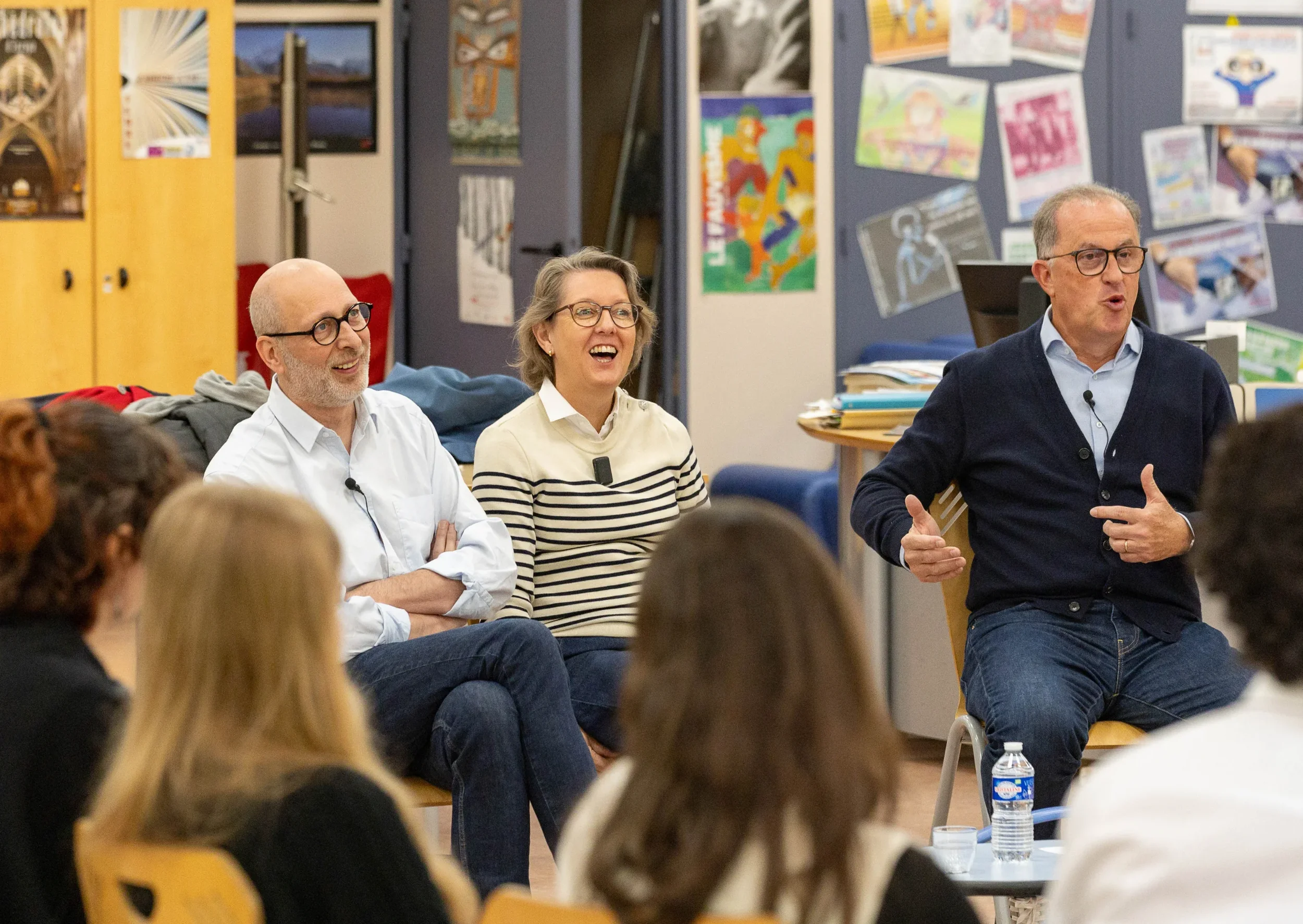 Trois personnes sont assises et une personne parle lors d'une réunion ou d'une discussion en intérieur avec des affiches colorées sur le mur derrière eux.