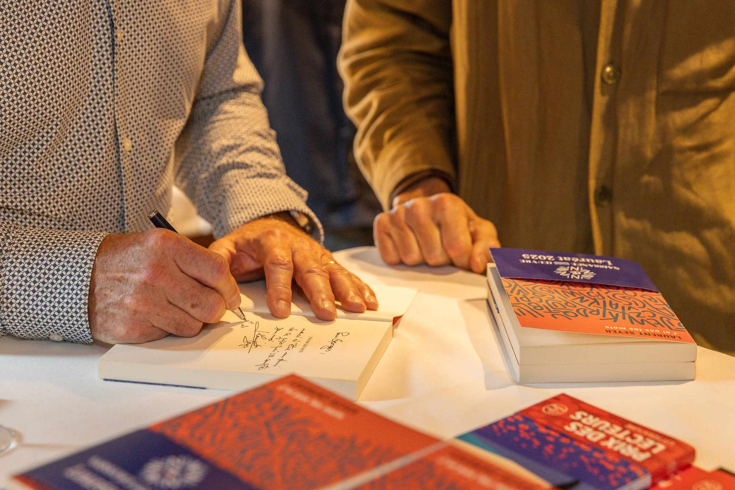 Deux personnes signant un livre lors d'une rencontre littéraire, avec des livres empilés sur la table.