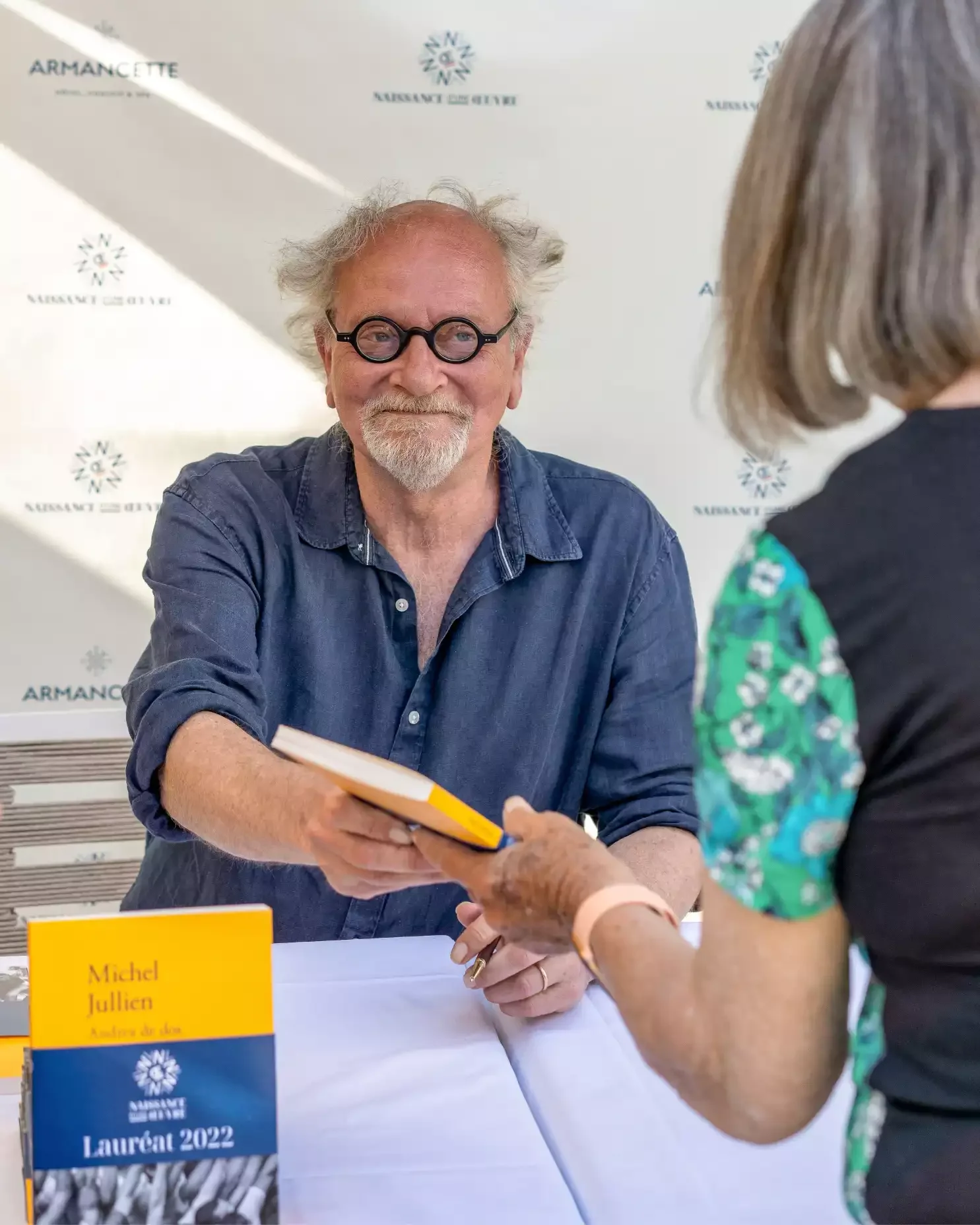 Un homme âgé avec des lunettes rondes, une barbe blanche, en chemise en denim, souriant en signant un livre pour une femme lors d'une signature de livre.