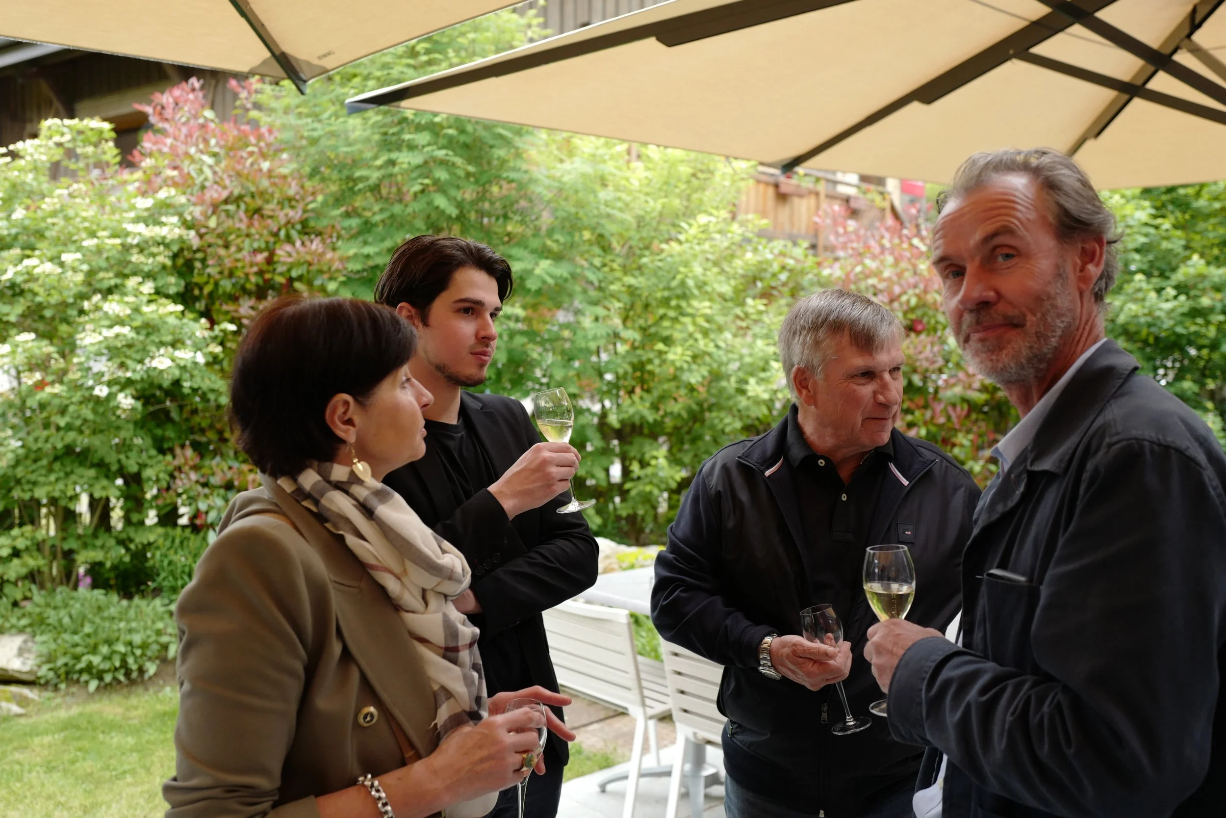 Groupe de personnes lors d'une réunion ou d'une célébration en extérieur, discutant avec des verres de vin ou de champagne, sous un grand parasol, avec un jardin fleuri en arrière-plan.