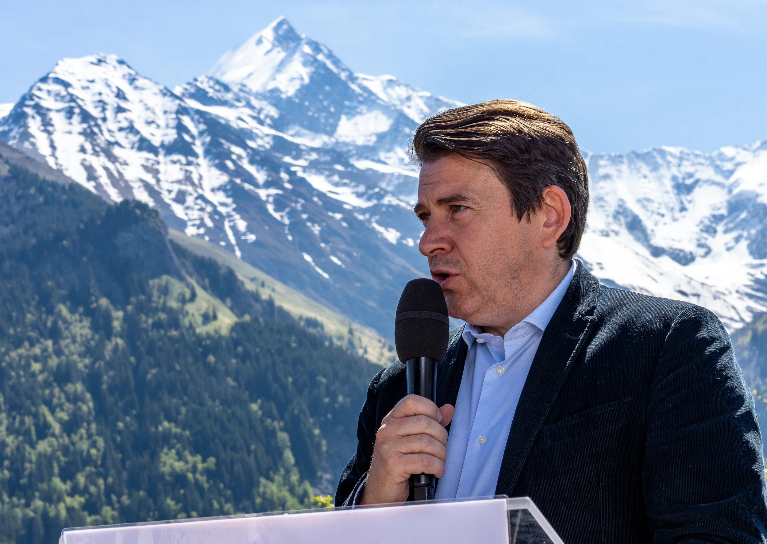 Un homme parlant dans un micro lors d'un discours en plein air avec des montagnes enneigées en arrière-plan.