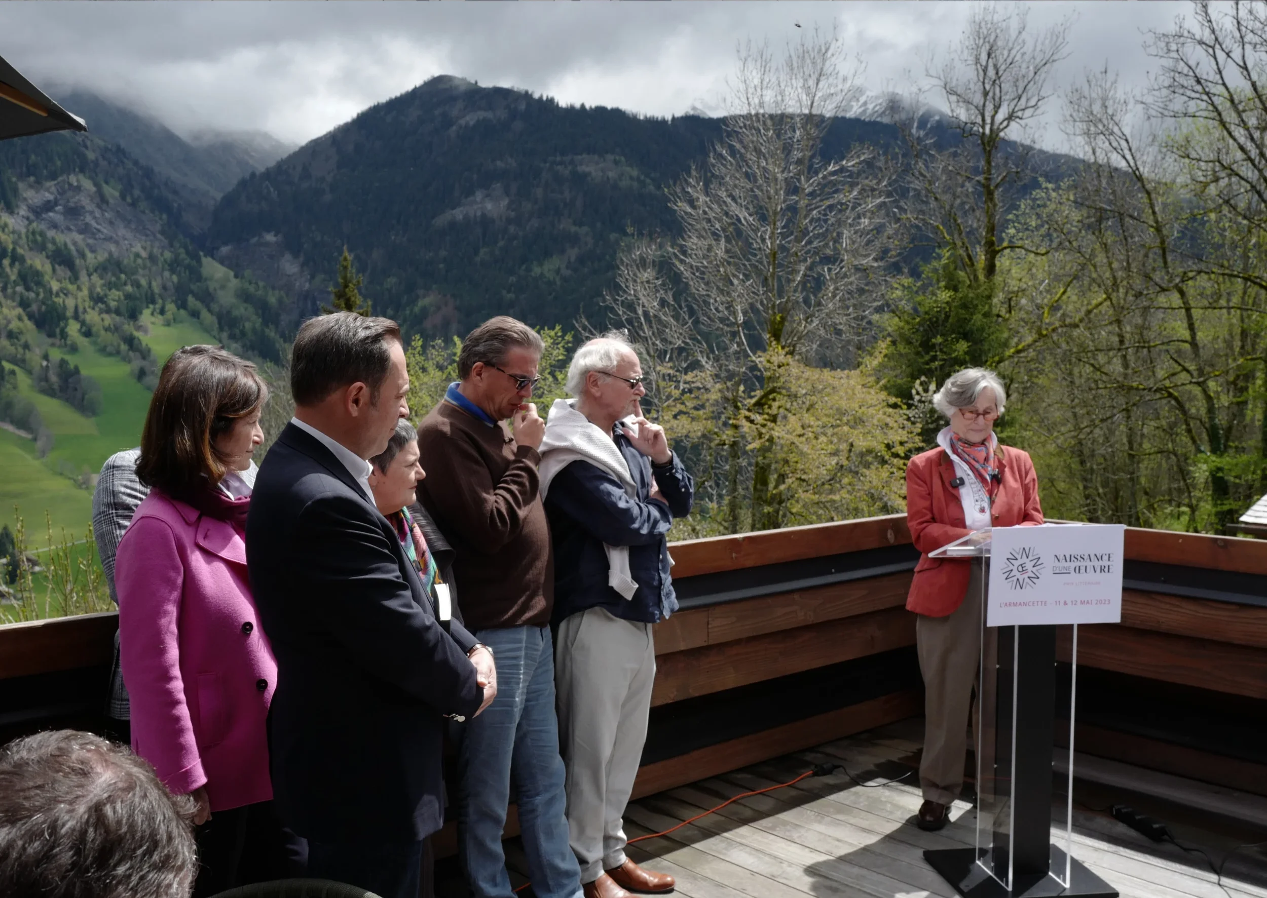Groupe de personnes debout en plein air contre un fond de montagnes, certaines ont les yeux fermés, et une femme à droite parle derrière un podium avec un panneau, lors d'une cérémonie ou d'un rassemblement.