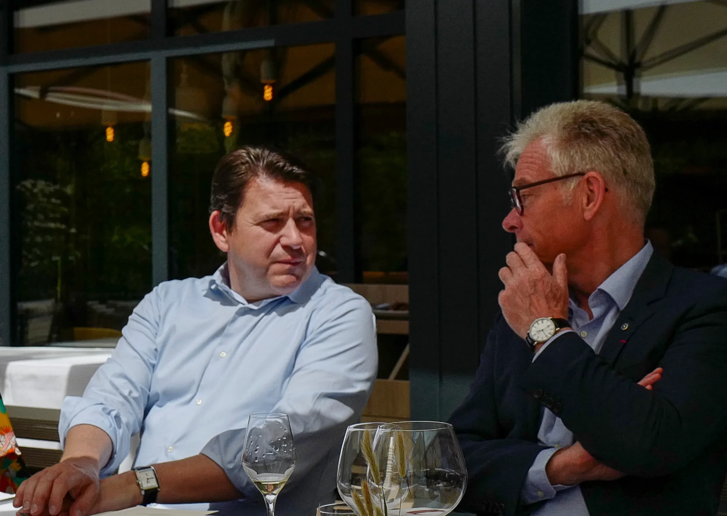 Deux hommes en conversation lors d'un dîner, assis à une table avec des verres de vin, dans un cadre intérieur.