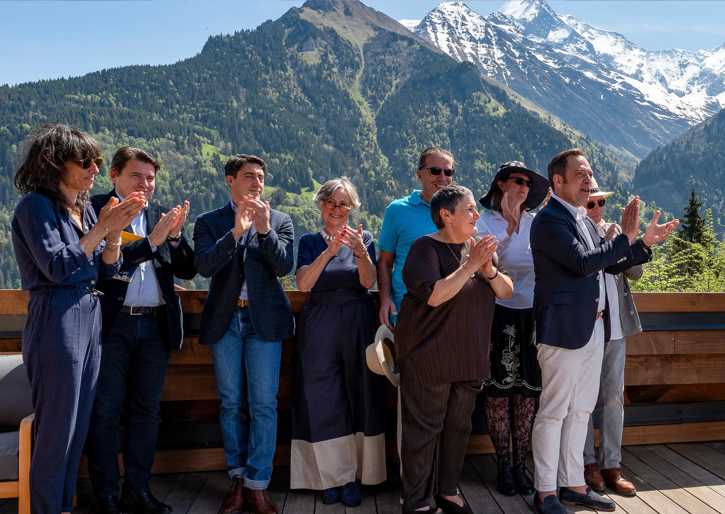 Groupe de huit personnes applaudissant lors d'un événement en plein air, avec des montagnes enneigées en arrière-plan.