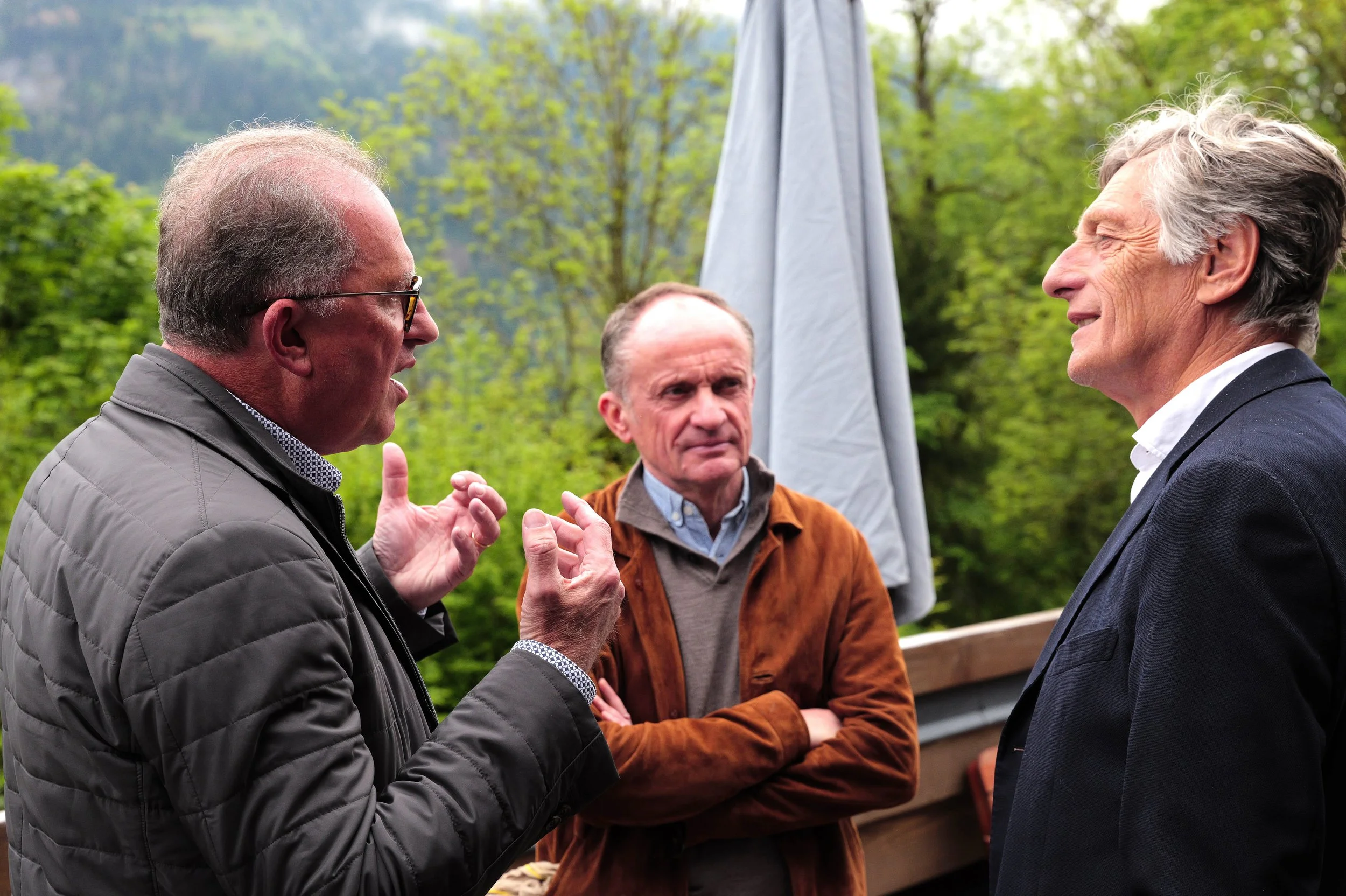 Trois hommes discutent en plein air, deux à l'avant et un en arrière-plan, avec des arbres verts et un ciel nuageux.