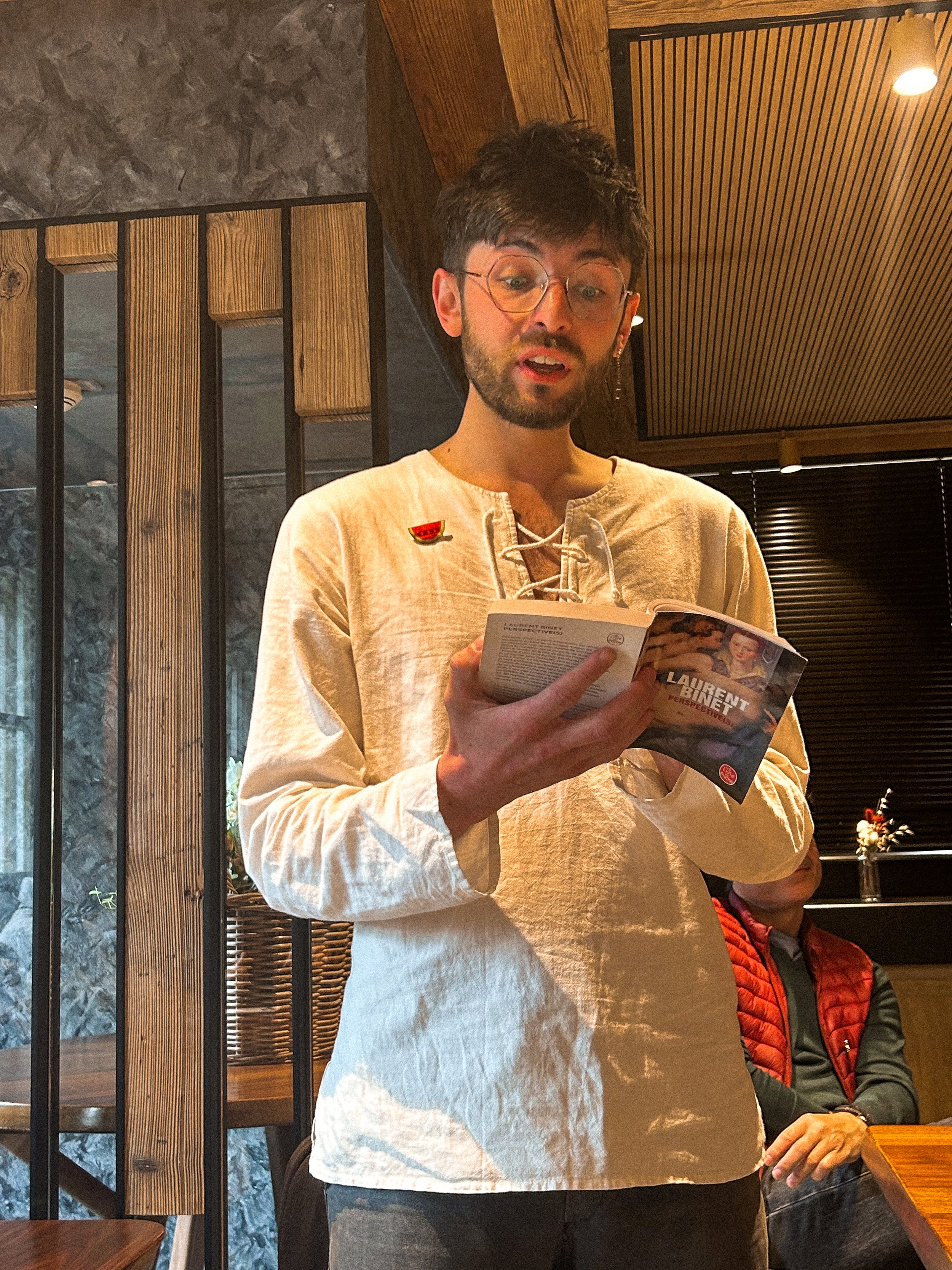 Une personne portant des lunettes, vêtue d'une chemise beige, tient une brochure ou un magazine dans un restaurant ou café avec un décor en bois et en pierre.