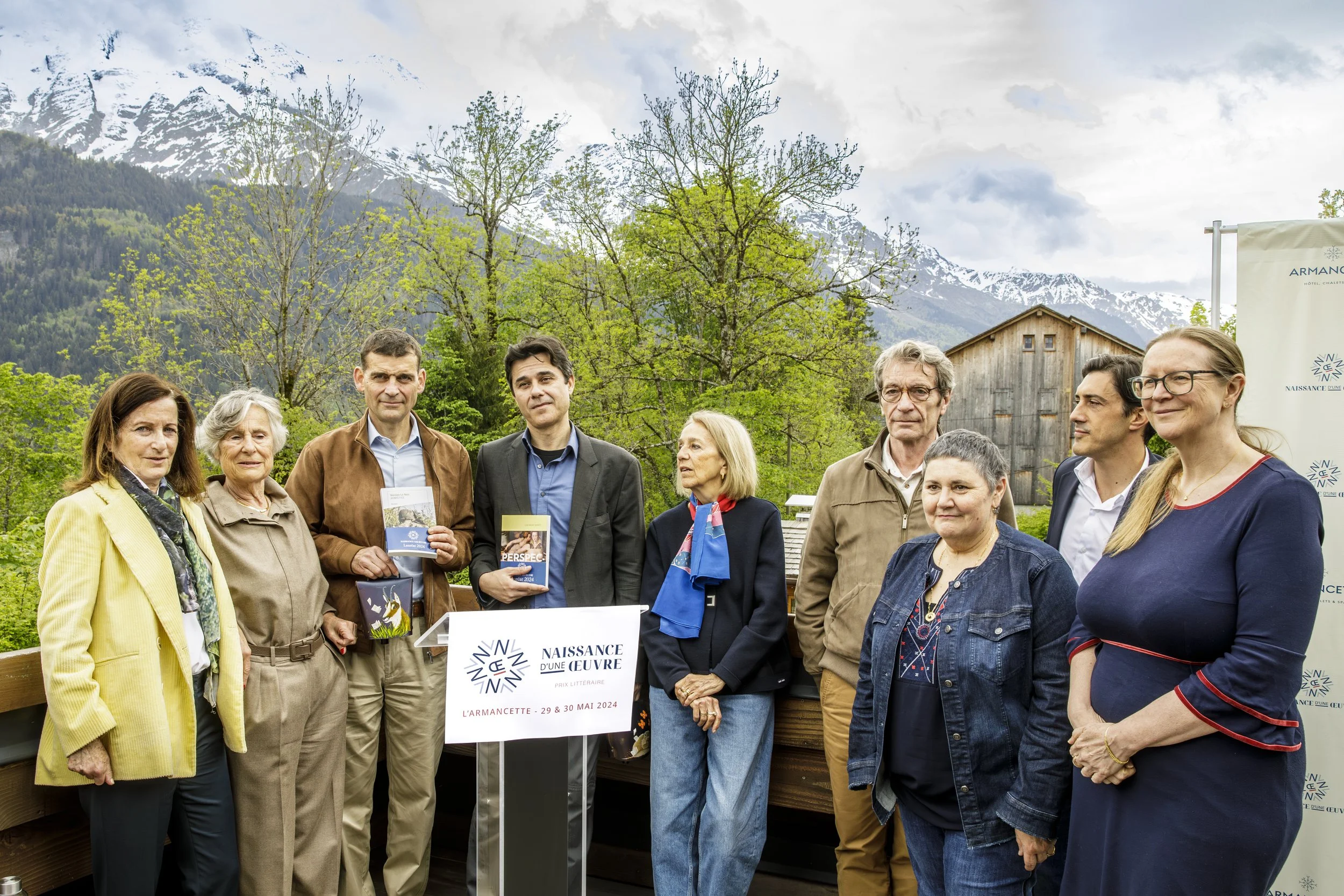 Groupe de personnes posant lors d'un événement en plein air avec montagnes enneigées en arrière-plan. Certains tiennent des livres et il y a une affiche indiquant la date 29-30 mai 2024, pour l'événement "L'ARMANCETTE".