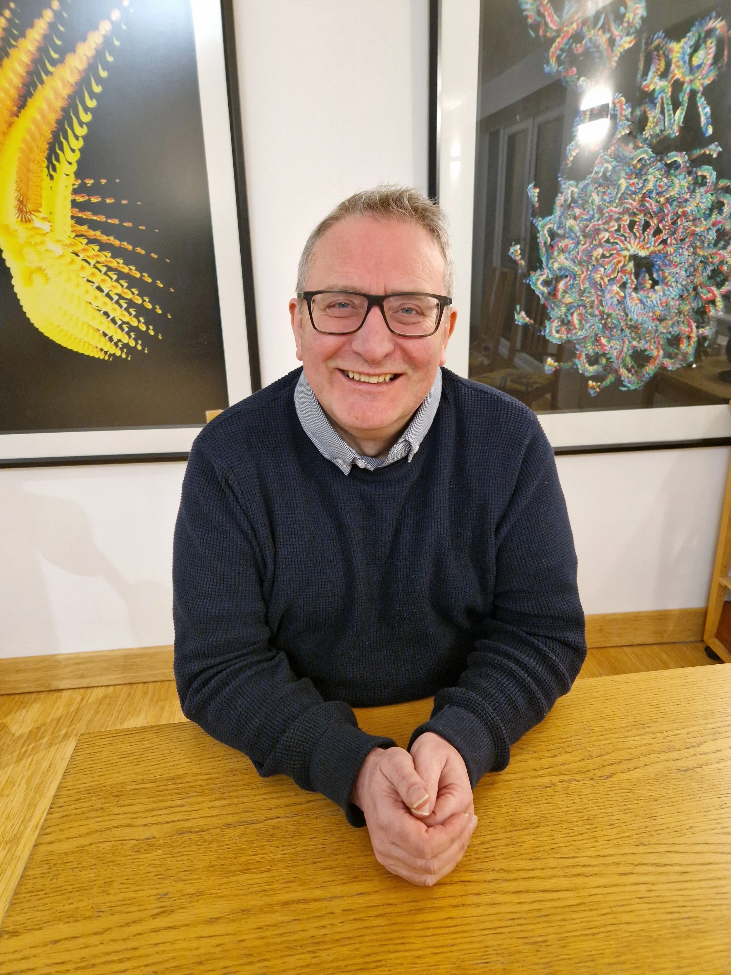 A smiling man, John Twining, with glasses sitting at a wooden table with his hands folded in front of him, with colourful artwork behind him.