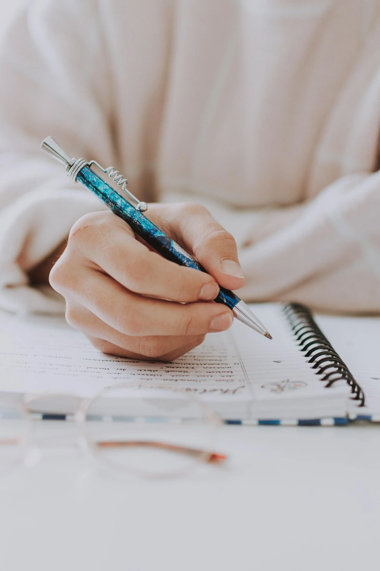 Close-up of a person writing in a spiral notebook with a blue and silver pen. The person is wearing a light-colored long-sleeve shirt, and the focus is on the hand and pen. The notebook is open on a white surface, with some text visible on the pages.