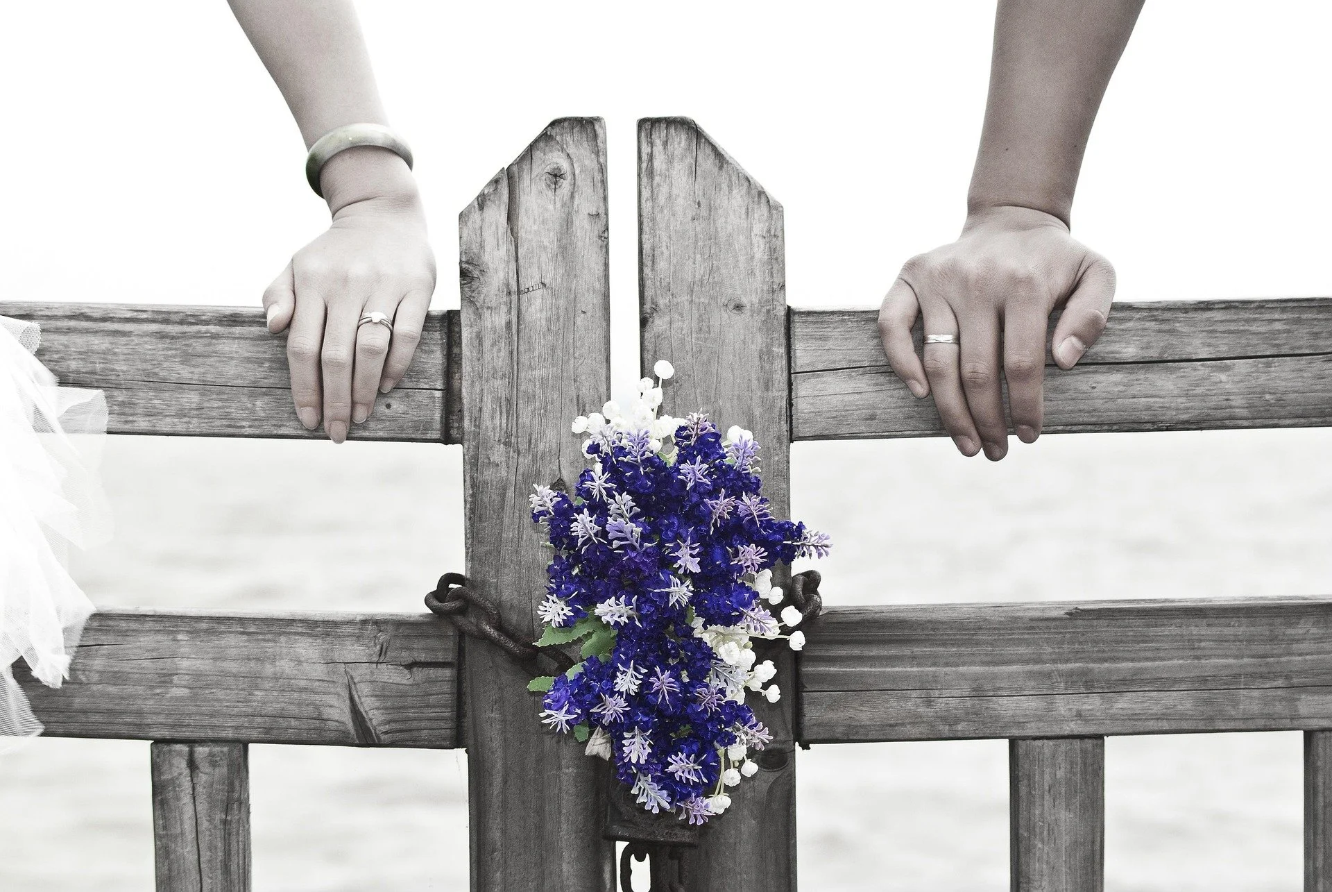 A pair of hands with rings and a bracelet hold the top of a wooden gate with a bunch of purple and white flowers hanging on the latch.