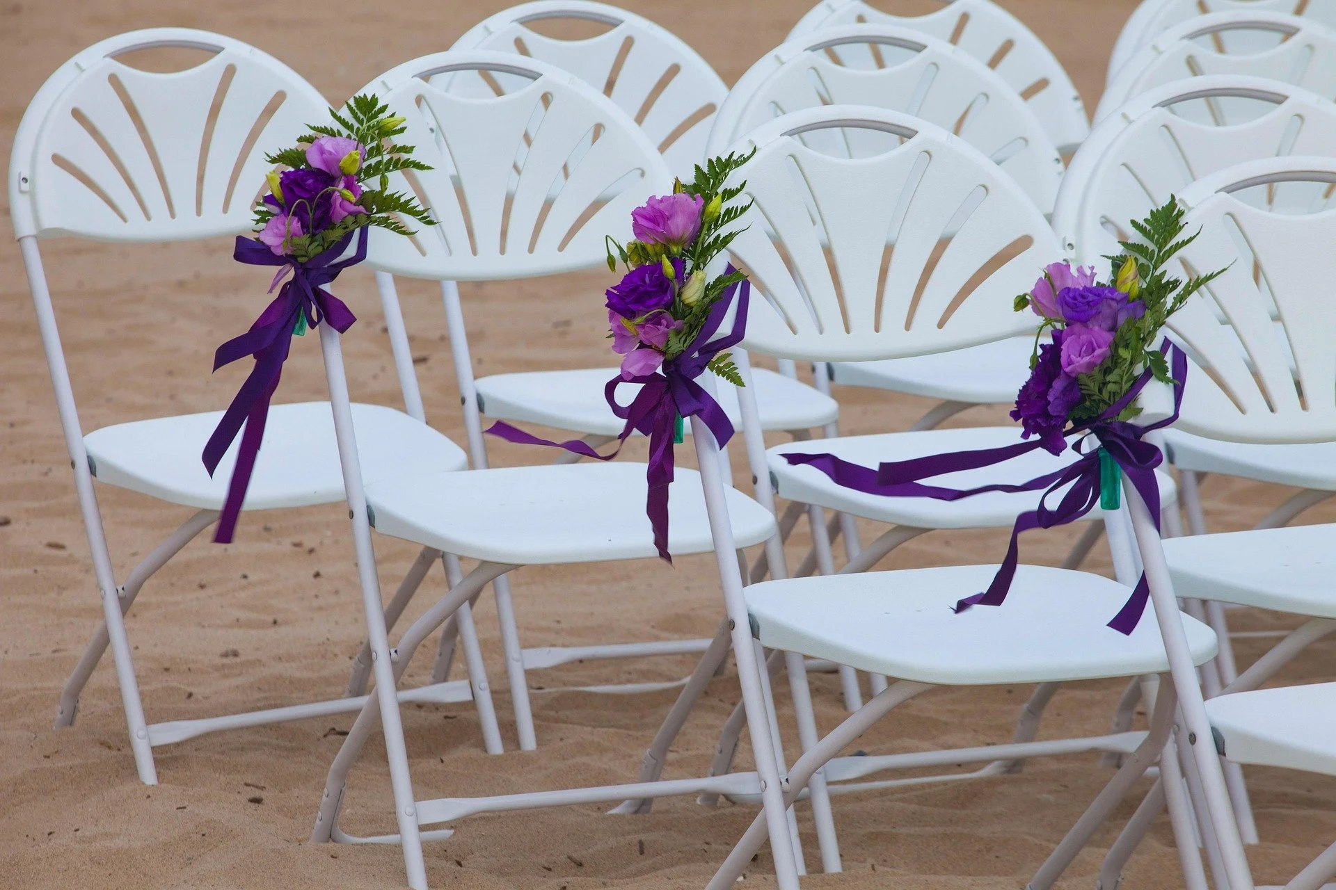 White folding chairs decorated with purple flowers and purple ribbons, arranged on a sandy surface for an outdoor wedding event.